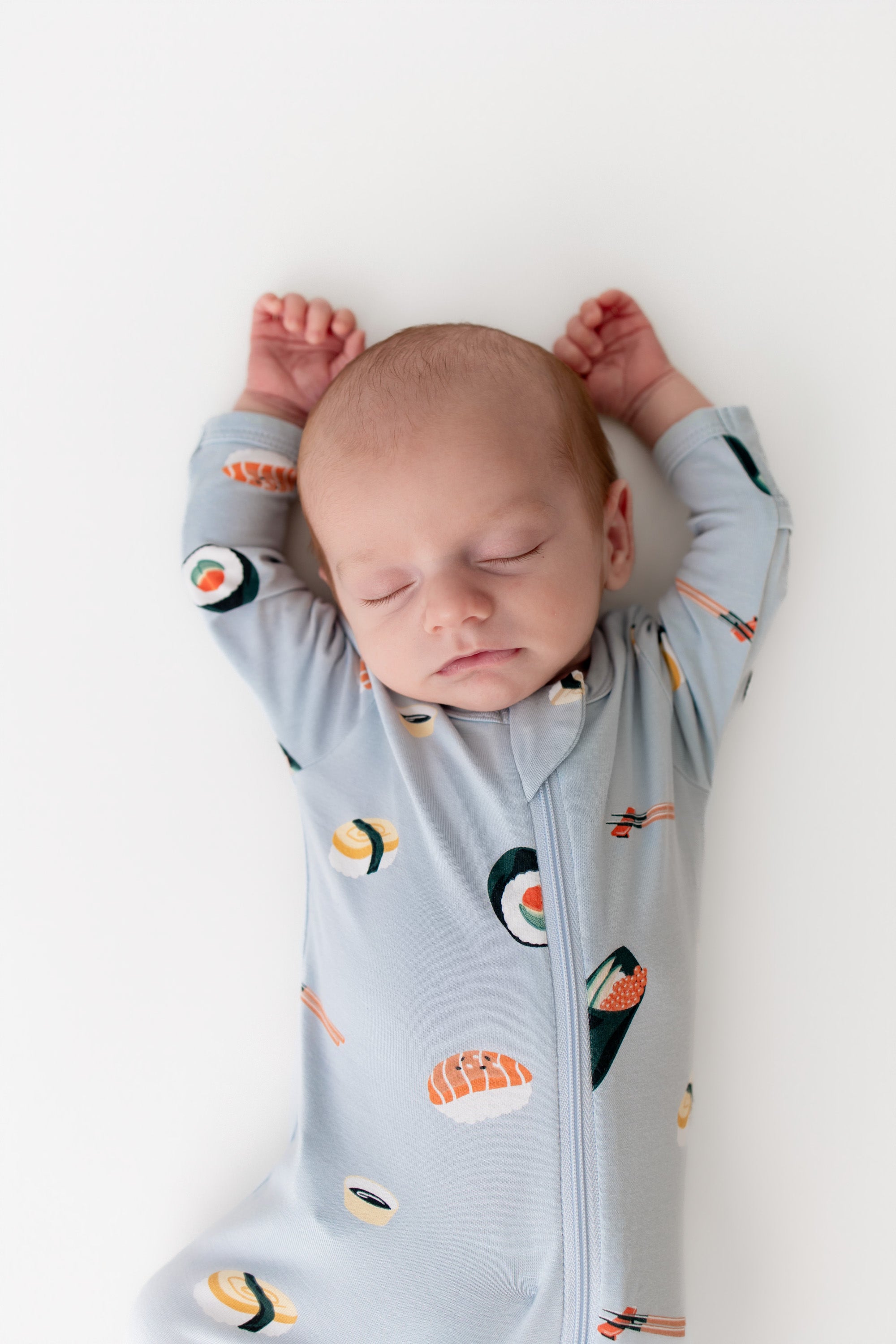 Baby sleeping with their arms up in ice sushi zipper footie on a white background