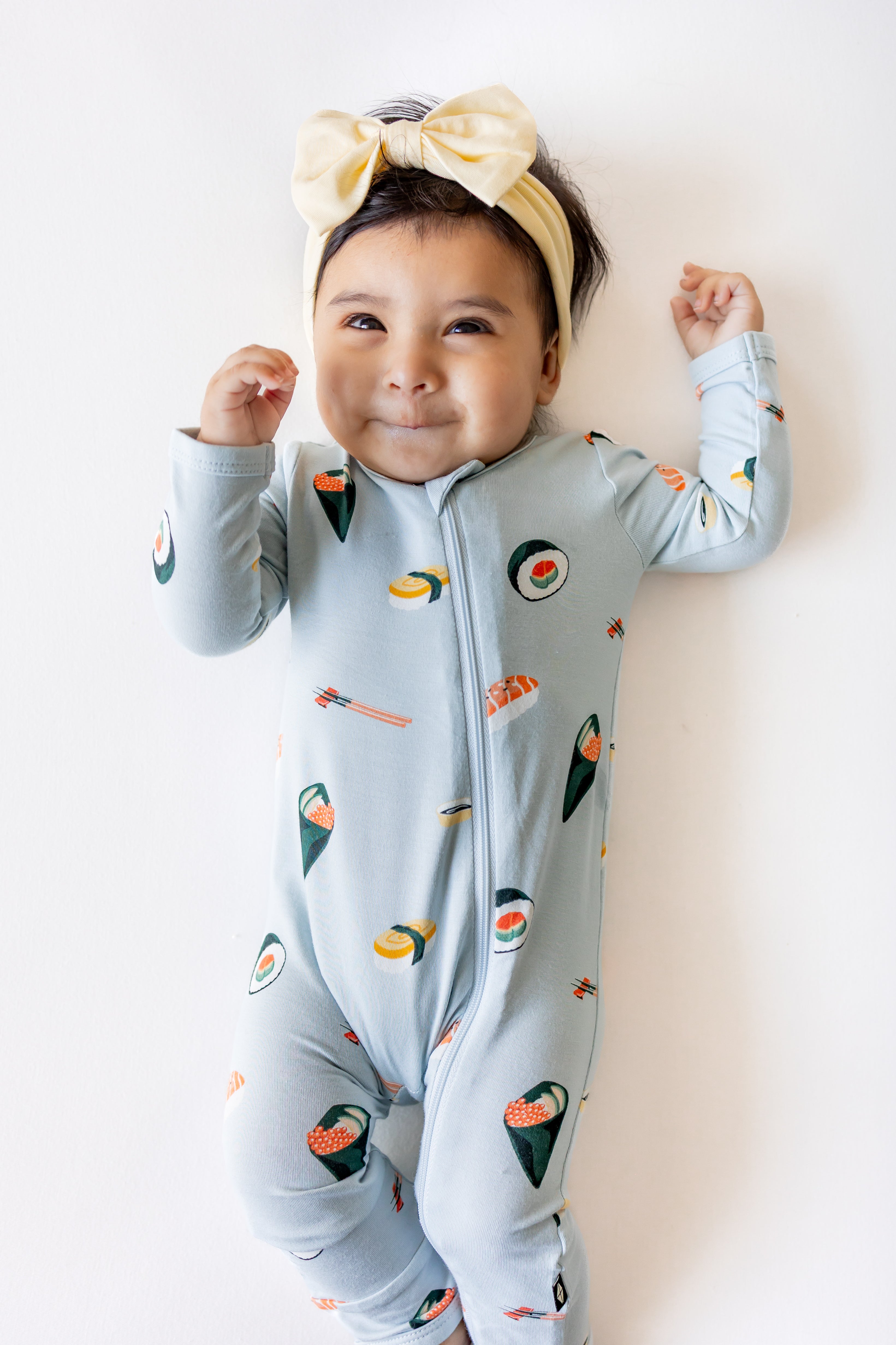 Baby laying down with a white background wearing ice sushi pajama set and a yellow bow