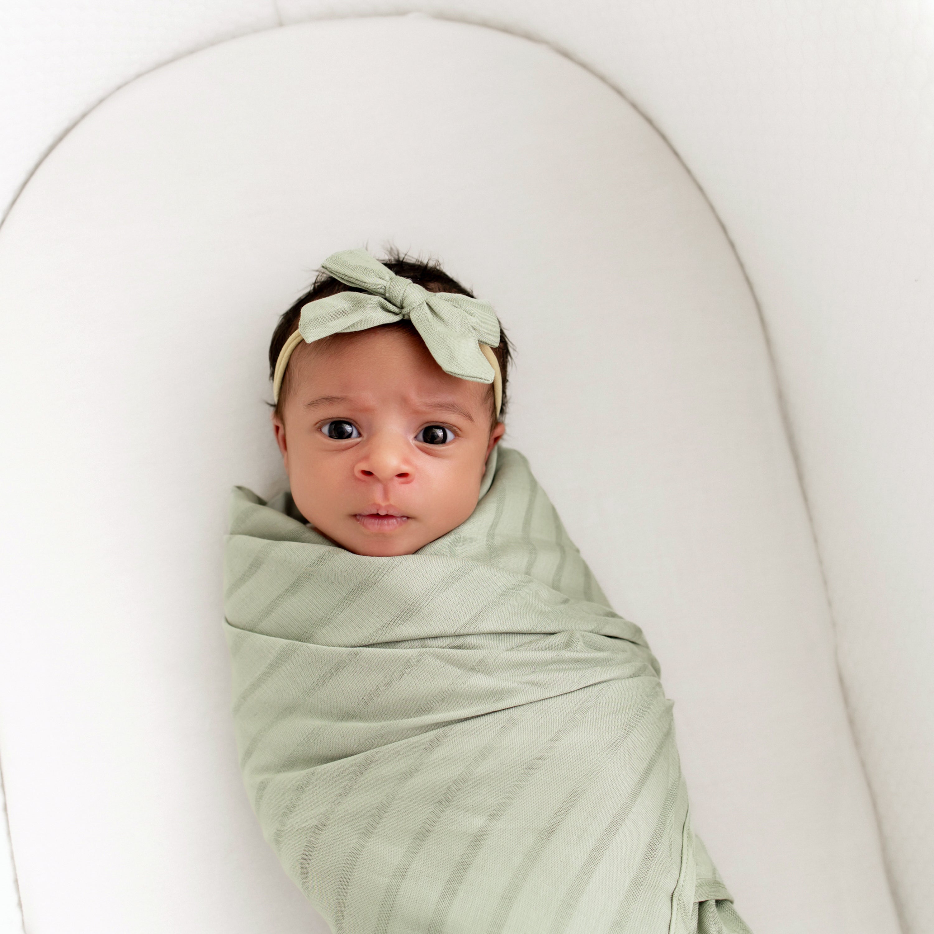 Baby swaddled in Bamboo Muslin Swaddle Blanket in Jojoba with matching bow headband