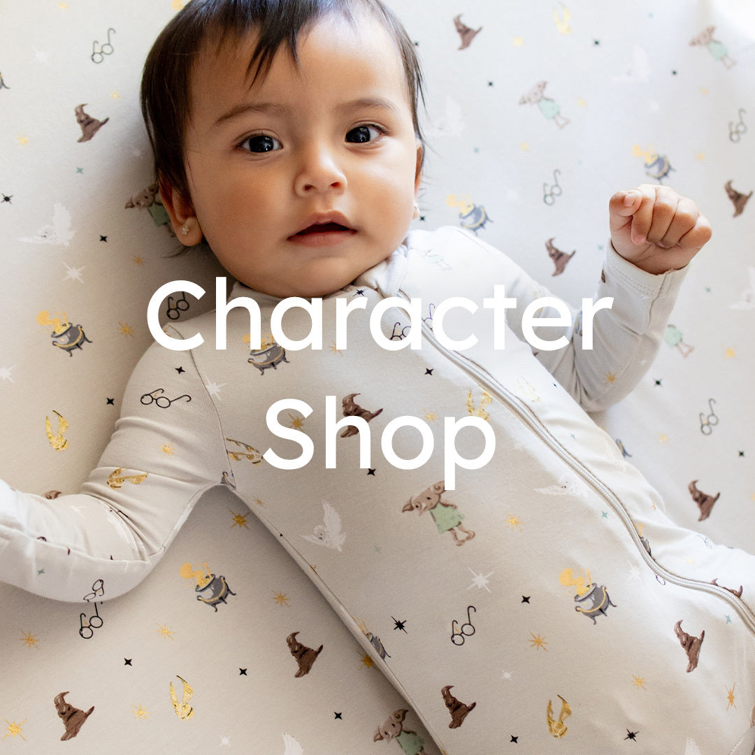 A baby lying on a patterned blanket wearing a long-sleeve zip-up sleeper. The sleeper has a cream base with small whimsical character prints in brown, yellow, and green.