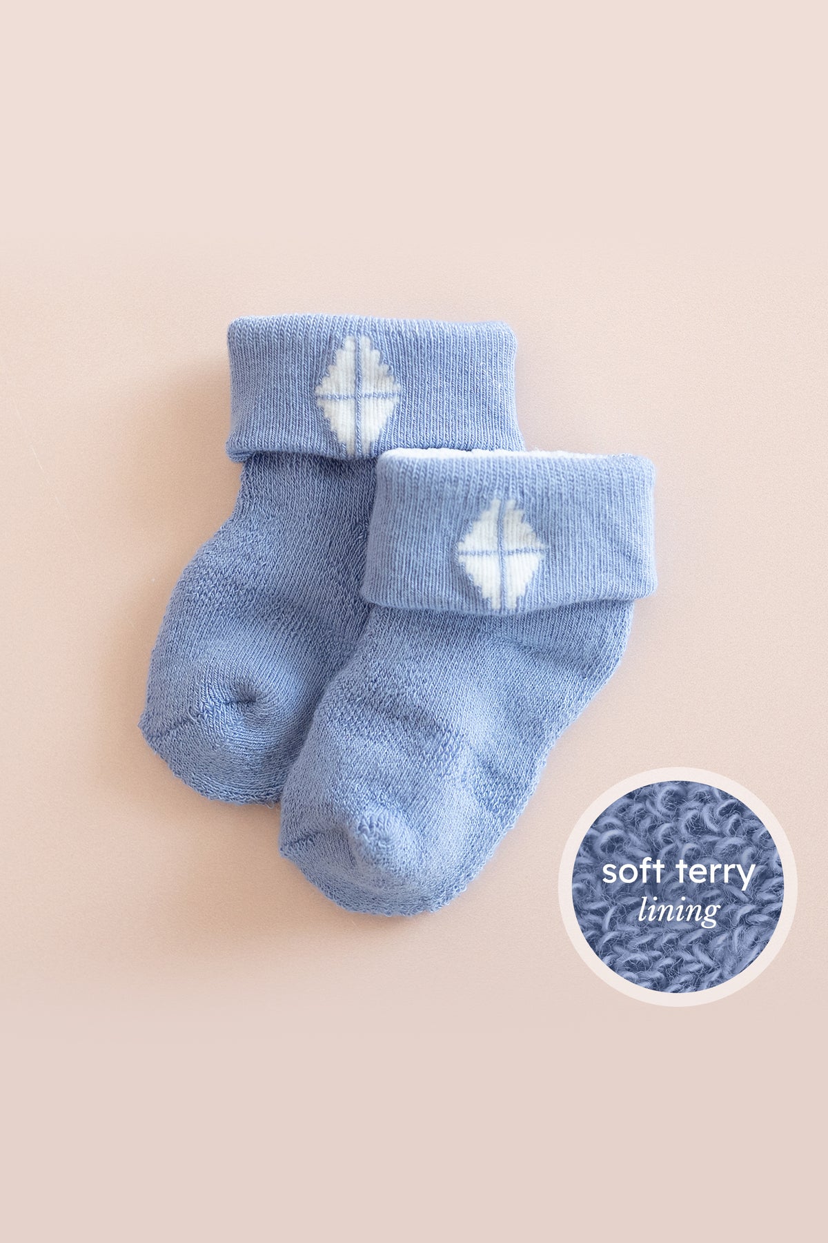 A pair of baby socks in a dusty light blue color, featuring a white embroidered snowflake design on the cuff. The socks are displayed on a soft beige background with a circular graphic overlay in the bottom right corner highlighting the 'soft terry lining'.