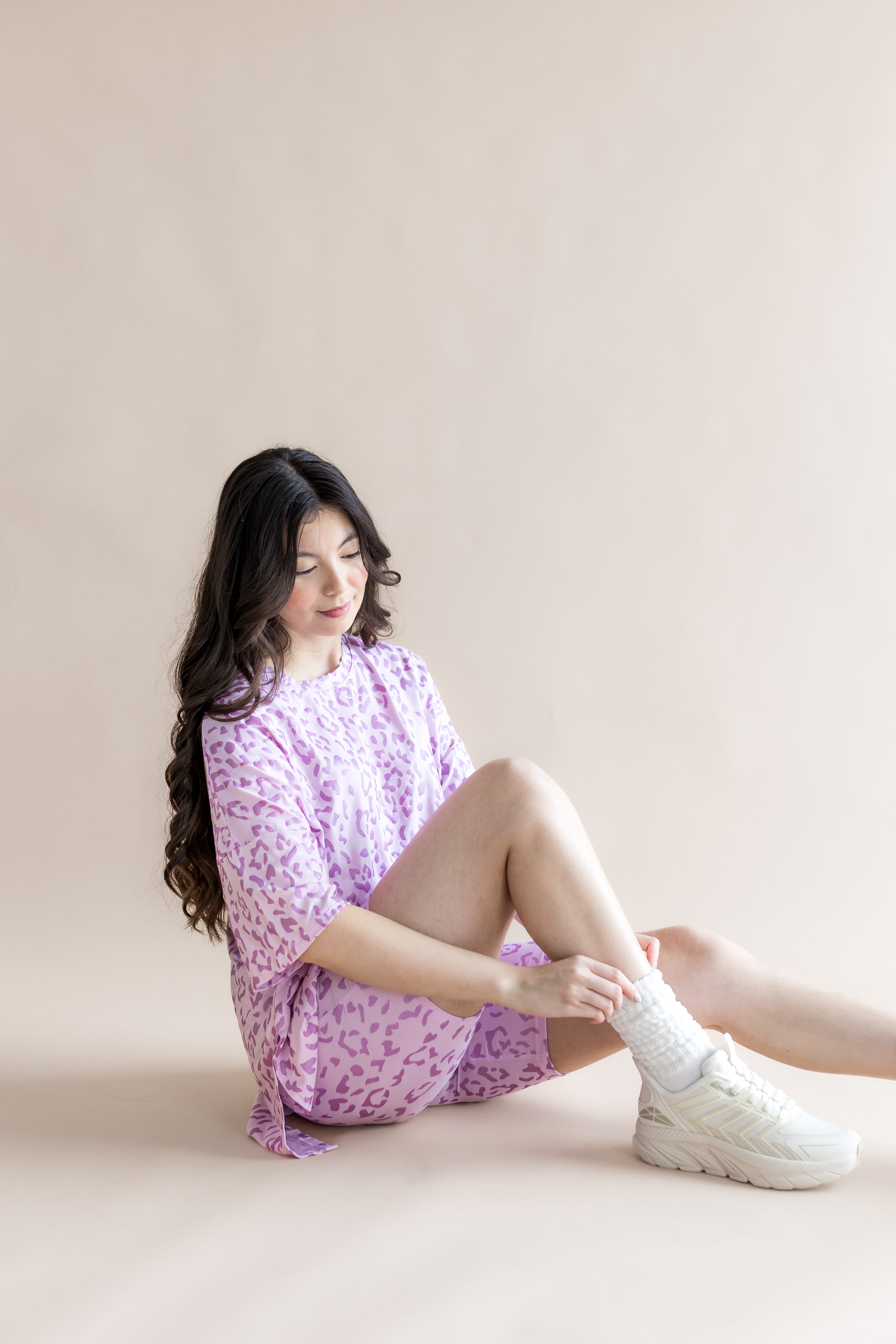 Model sitting in Women’s Biker Short Set in Thistle Leopard print with white socks and shoes on a beige background