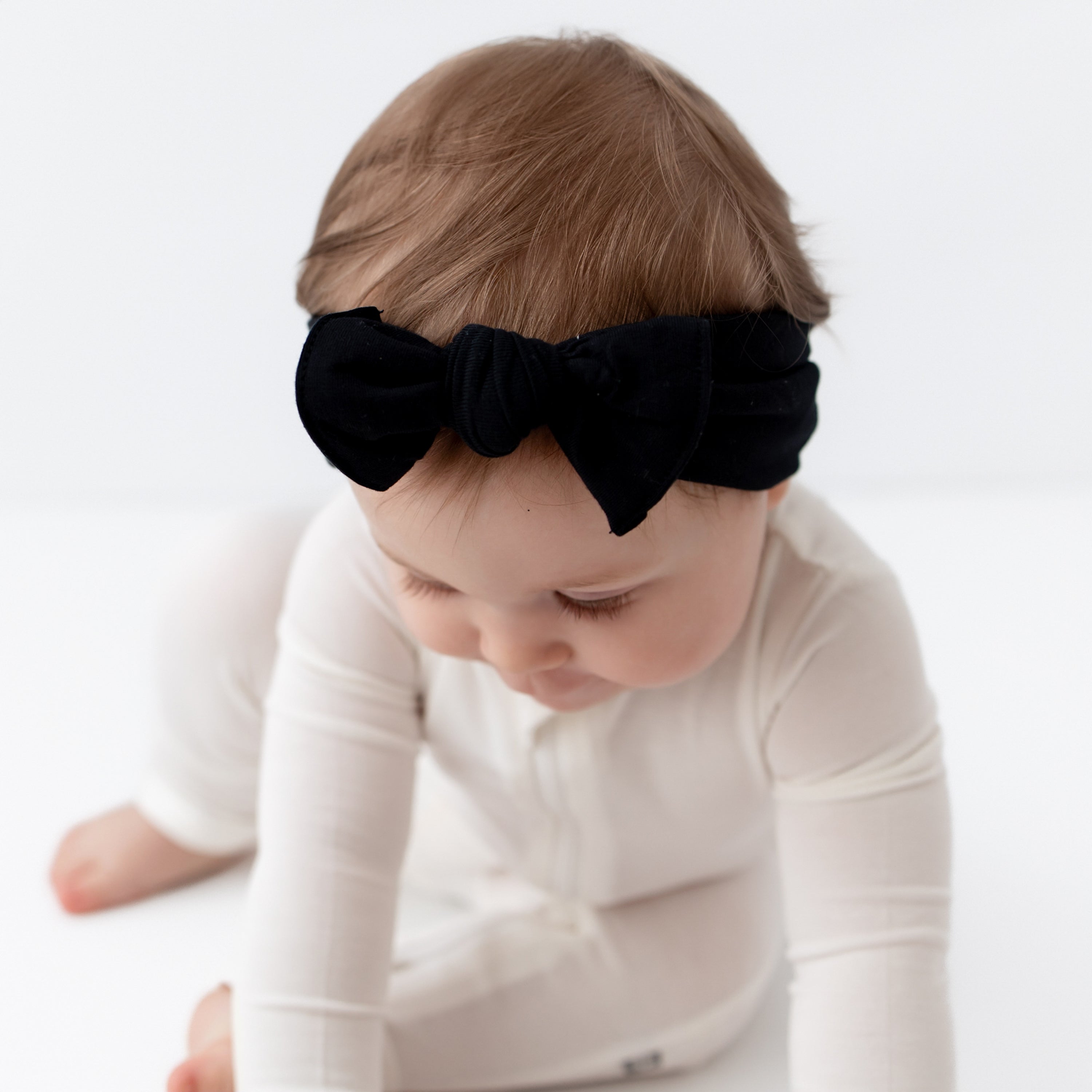 Close up of Knotted Bow Headband in Midnight on baby's head who is looking down and zippered romper in oat