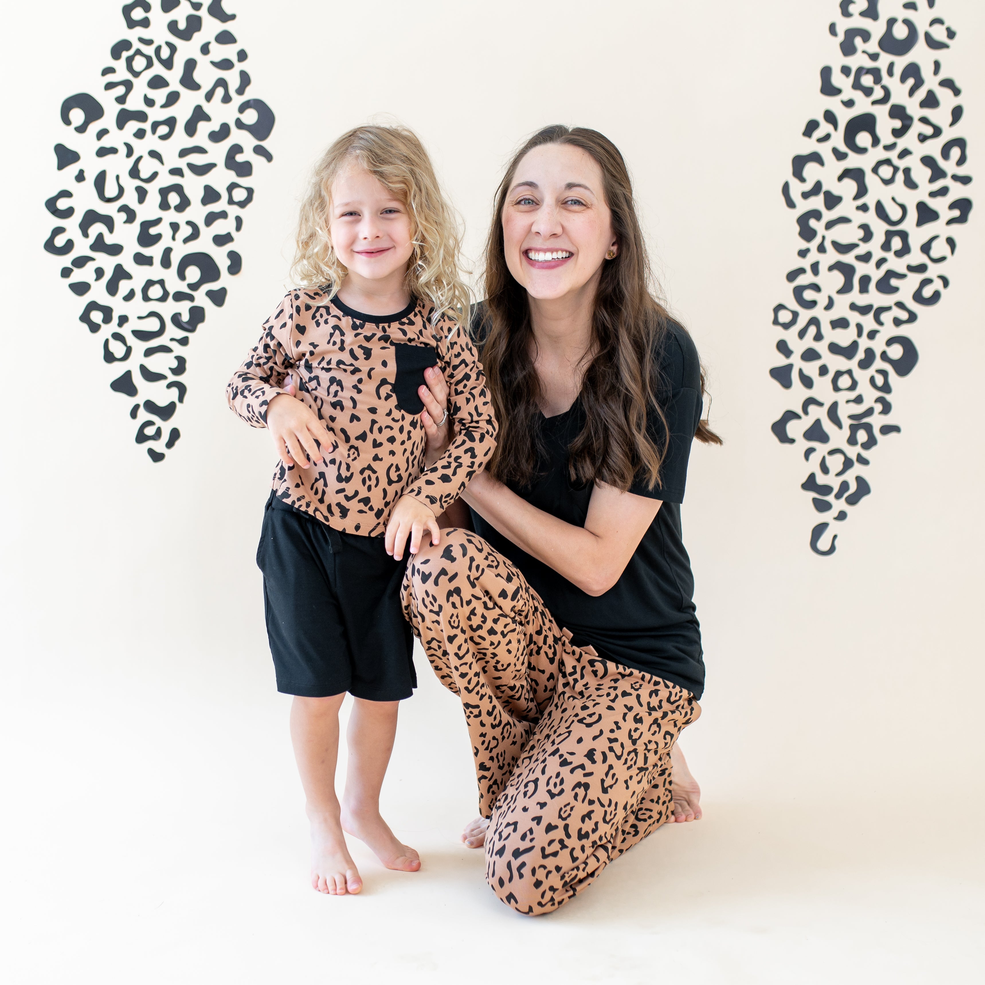 Mother kneeling beside her child both matching in Latte Leopard items in front of a leopard print backdrop