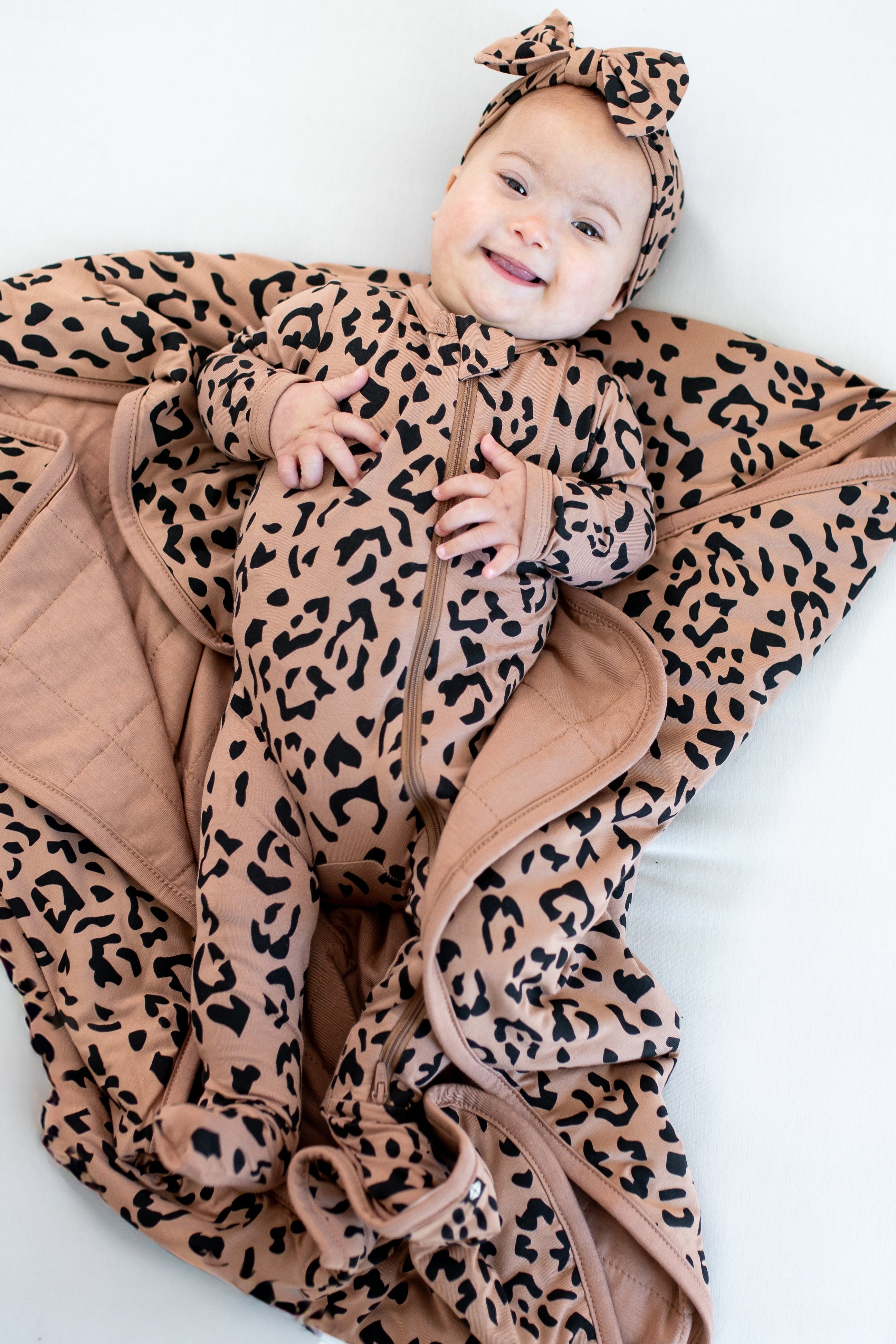 Infant laying on Baby Blanket in Latte Leopard wearing matching zippered footie pajamas and bow headband