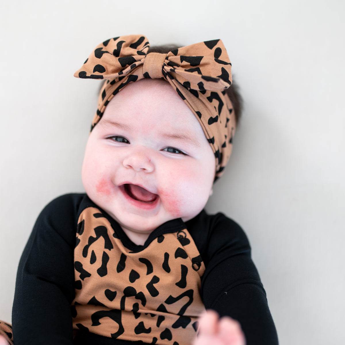 Smiling infant laying on a light colored blanket wearing the Bow Headband in Latte Leopard and matching longall