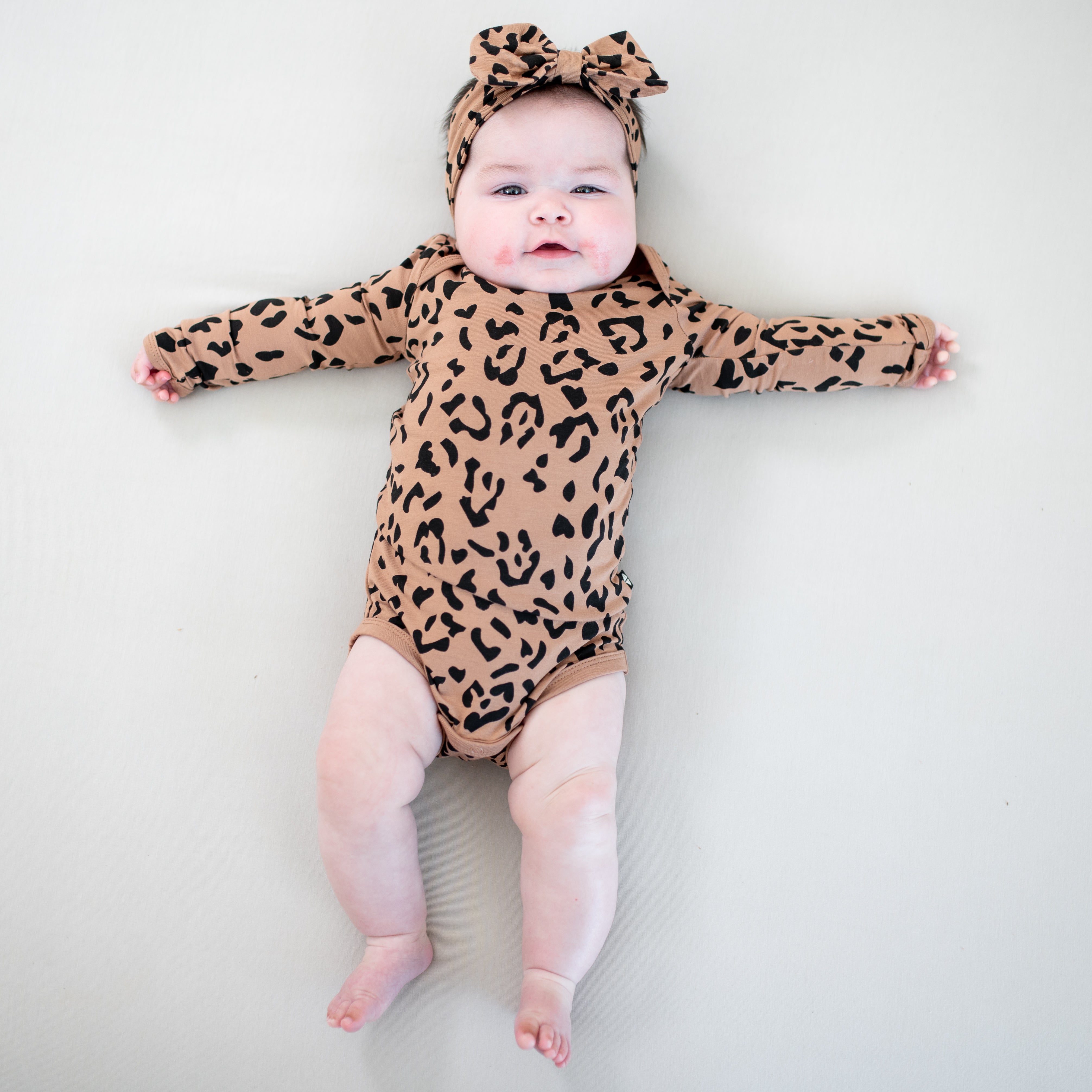 Infant laying on a light colored blanket with her arms outstretched wearing the Long Sleeve Bodysuit in Latte Leopard and matching bow headband