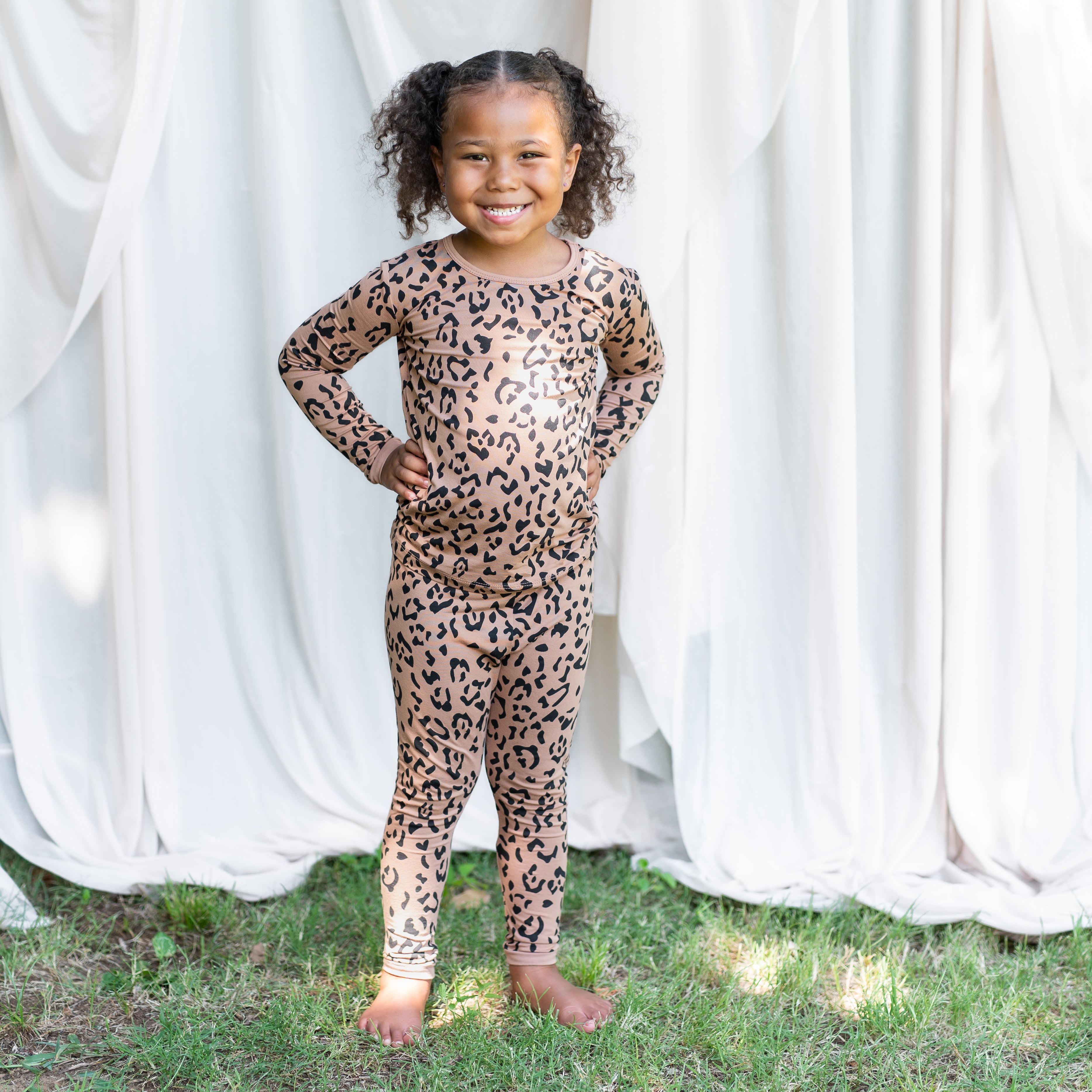 Smiling young girl standing in the grass with her hands on her hips wearing the Long Sleeve Pajamas in Latte Leopard