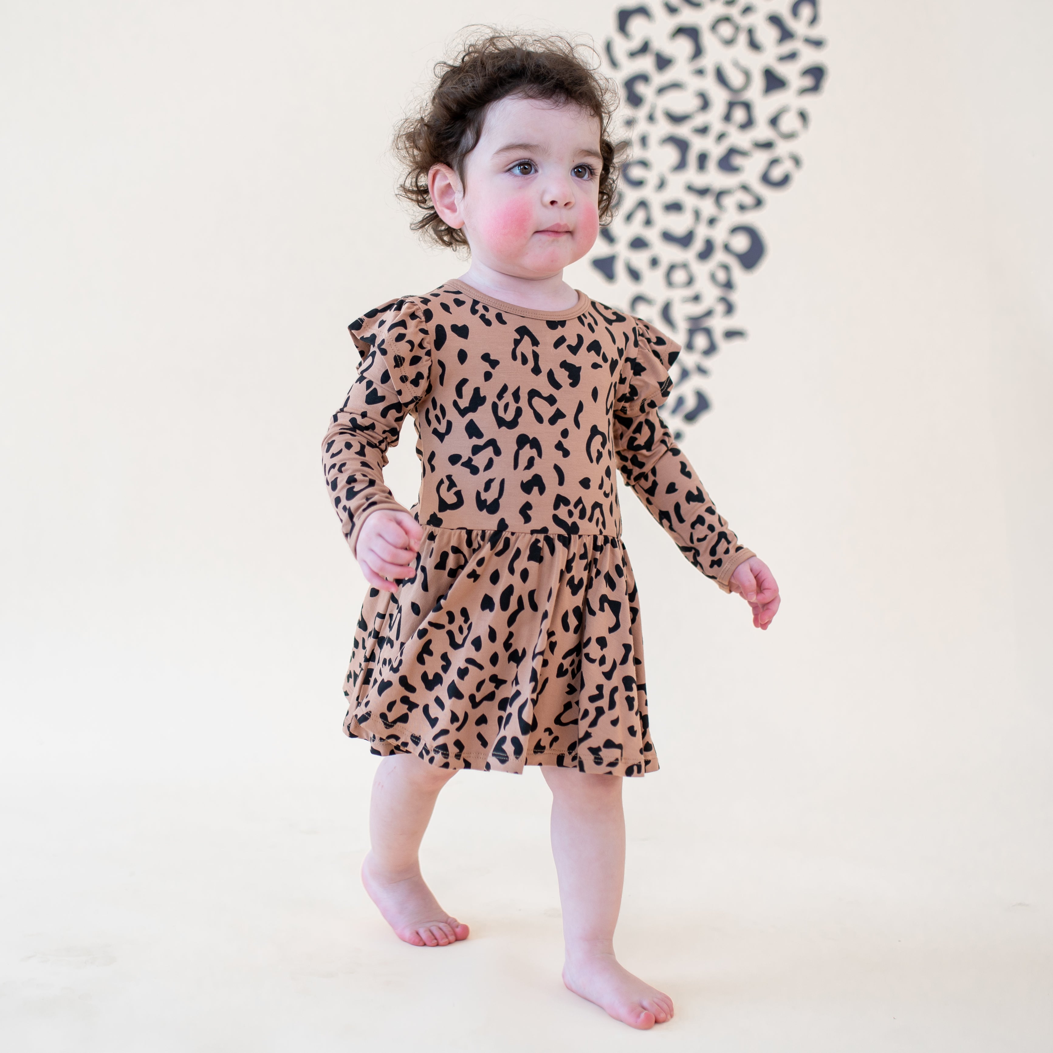 Toddler walking wearing the Long Sleeve Twirl Bodysuit Dress in Latte Leopard in front of a light beige leopard print backdrop