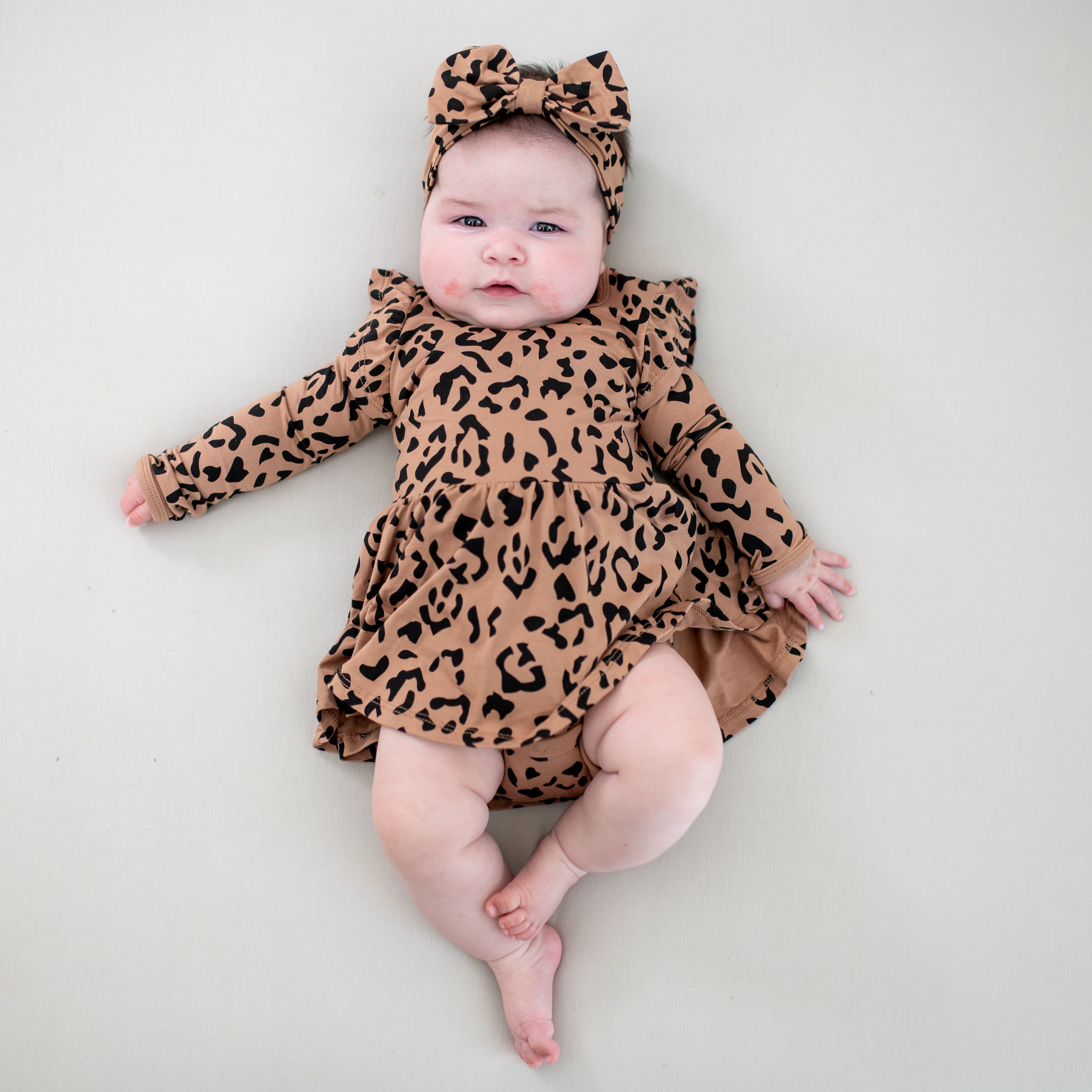 Infant laying on a light colored blanket wearing the Bow Headband in Latte Leopard and matching long sleeve twirl bodysuit dress