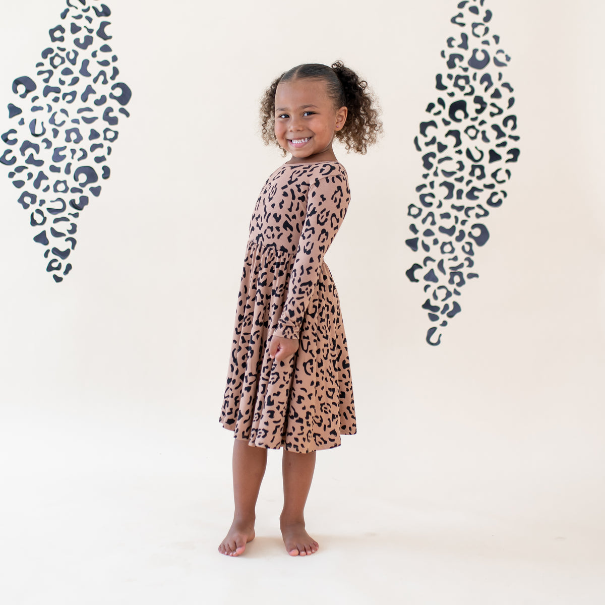 Young girl smiling standing wearing the Long Sleeve Twirl Dress in Latte Leopard in front of a light colored leopard print backdrop