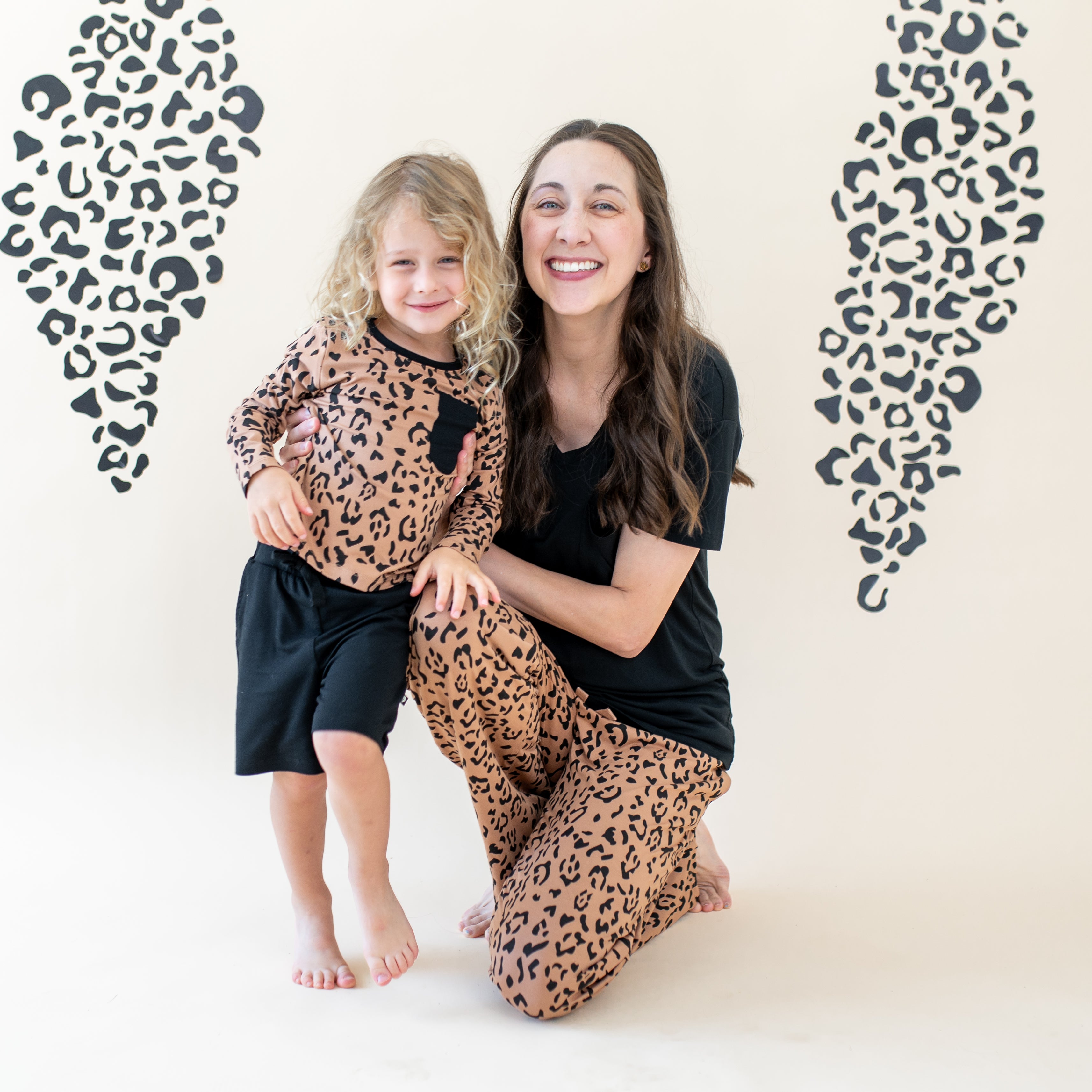 Mother wearing the Midnight women's V-neck and Latte leopard print lounge pants kneeling beside her child wearing the Long Sleeve Toddler Crew Neck Tee in Latte Leopard and Midnight drawstring shorts in front of a leopard print backdrop