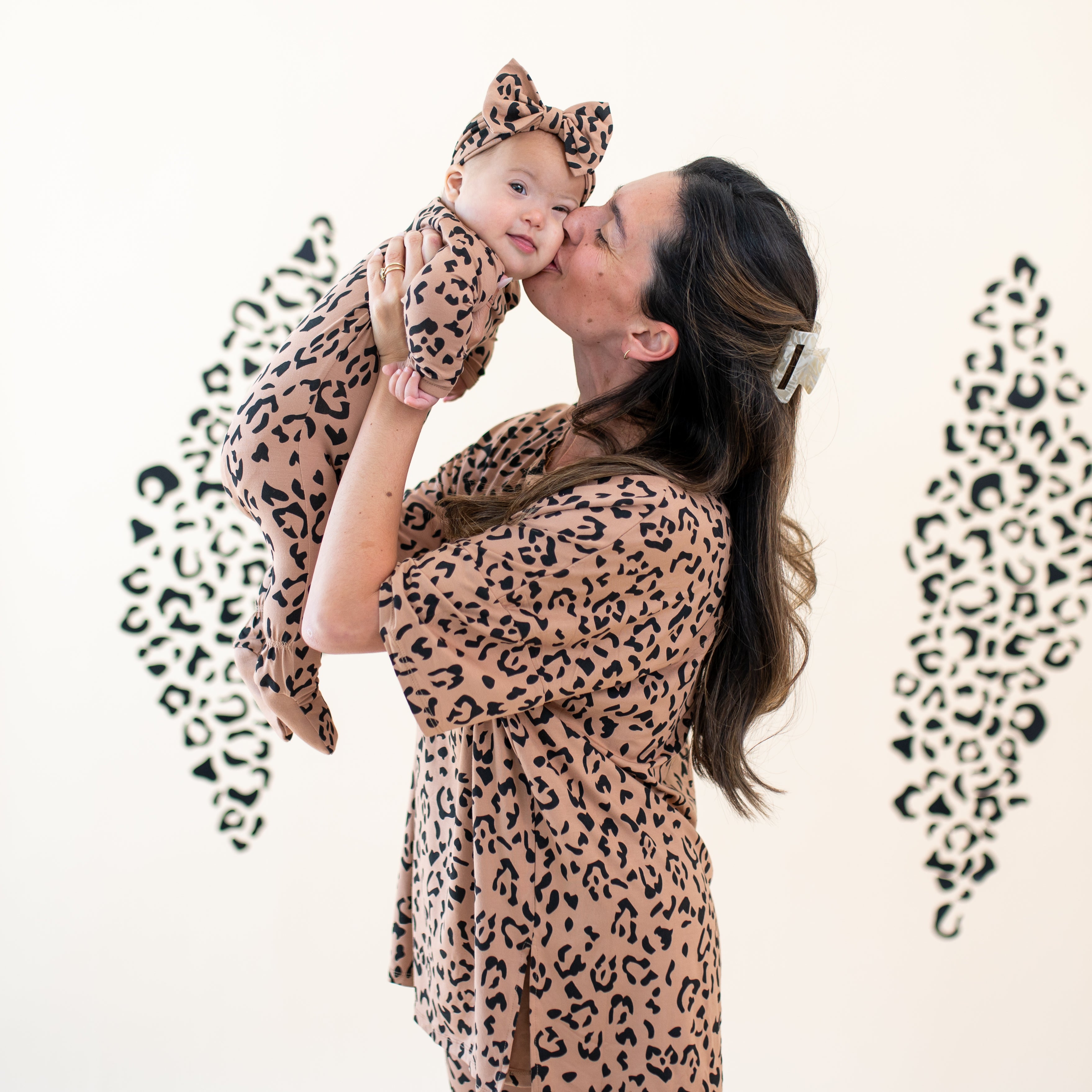 Mother wearing the Women's Biker Short Set in Latte Leopard holding her daughter in the air kissing her daughter on the cheek who is matching in the Bow Headband in Latte Leopard and matching zippered footie