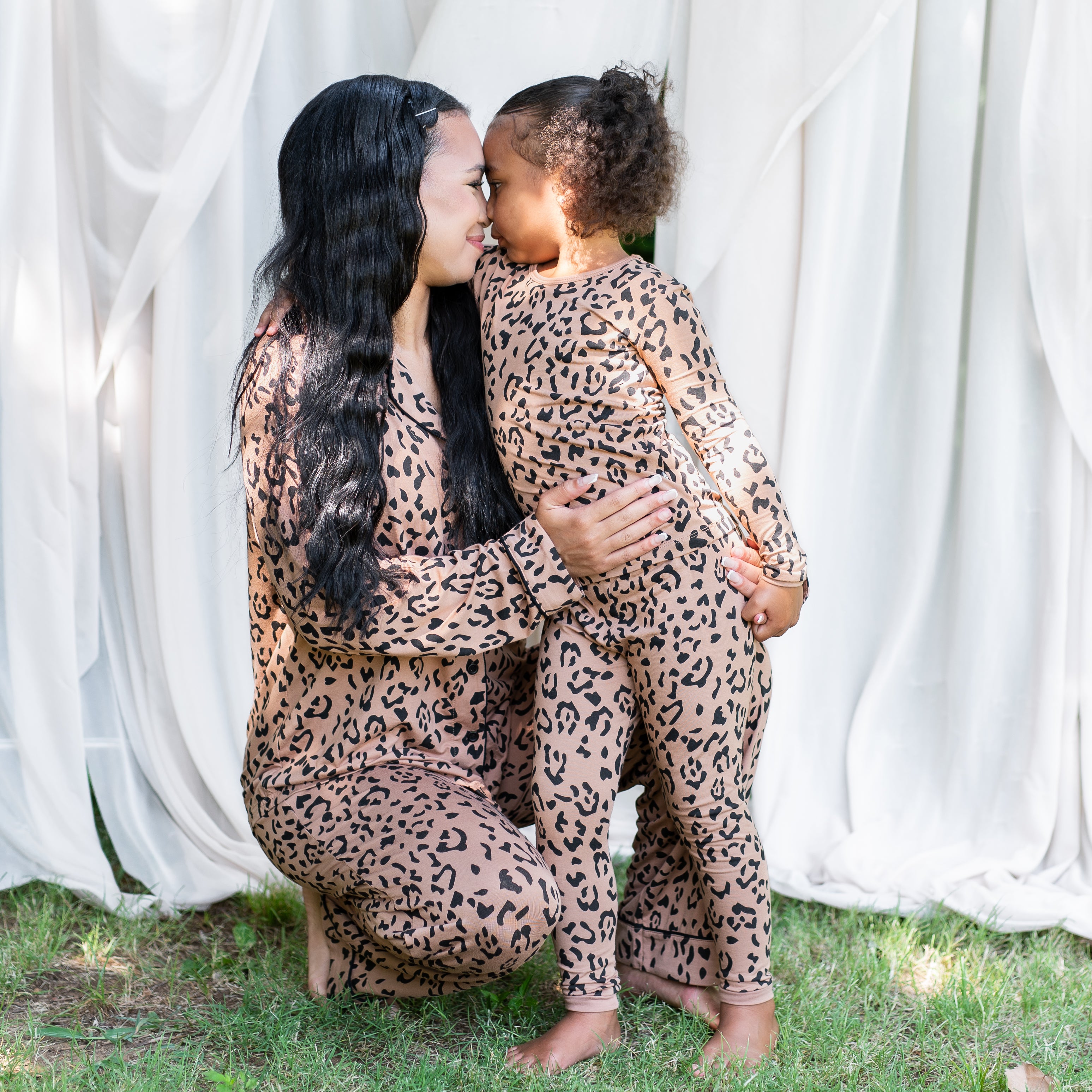 Mother kneeling beside her daughter in the grass with her arms around her both matching in Latte Leopard