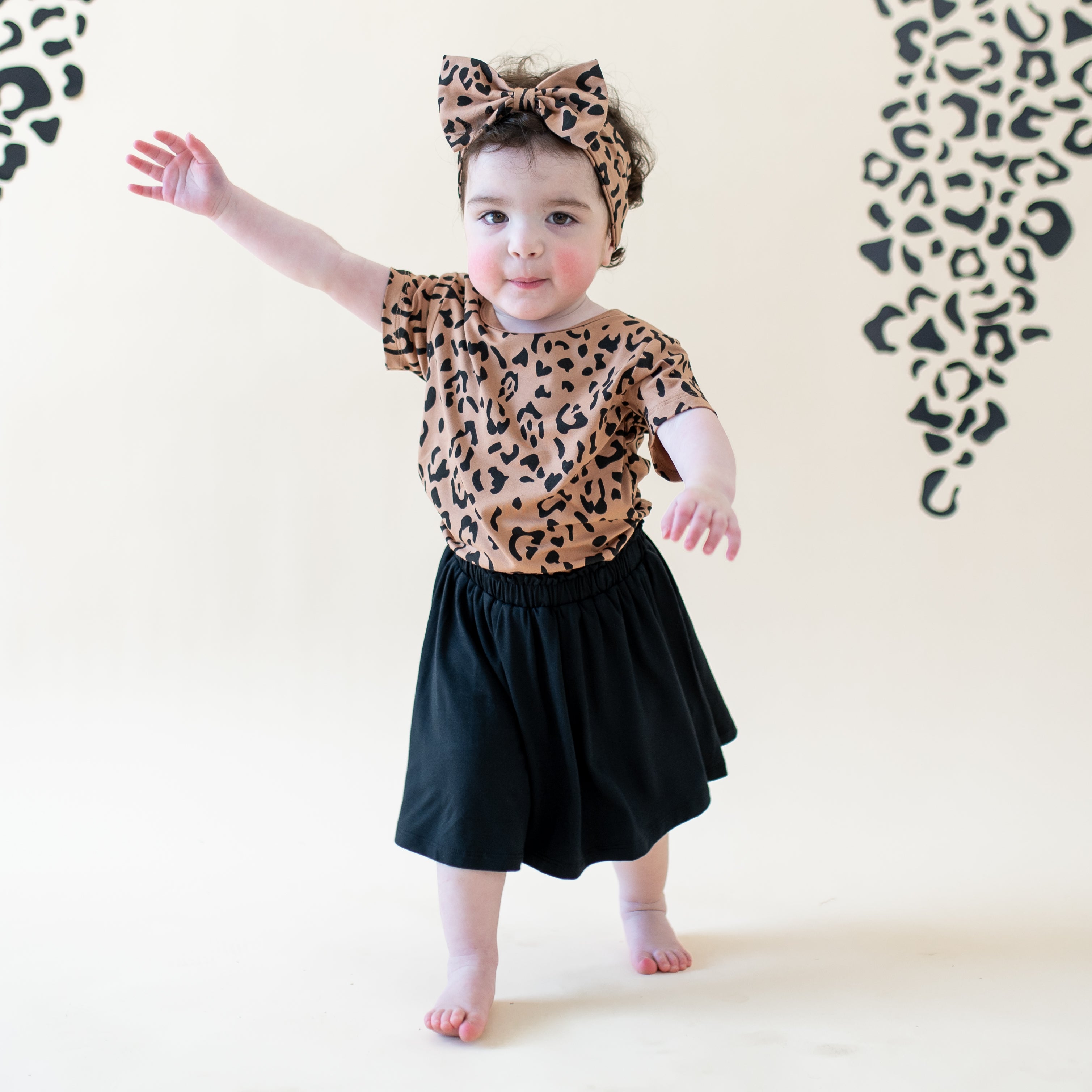 Toddler walking wearing the Toddler Basic Tee in Latte Leopard with matching bow headband and midnight skort
