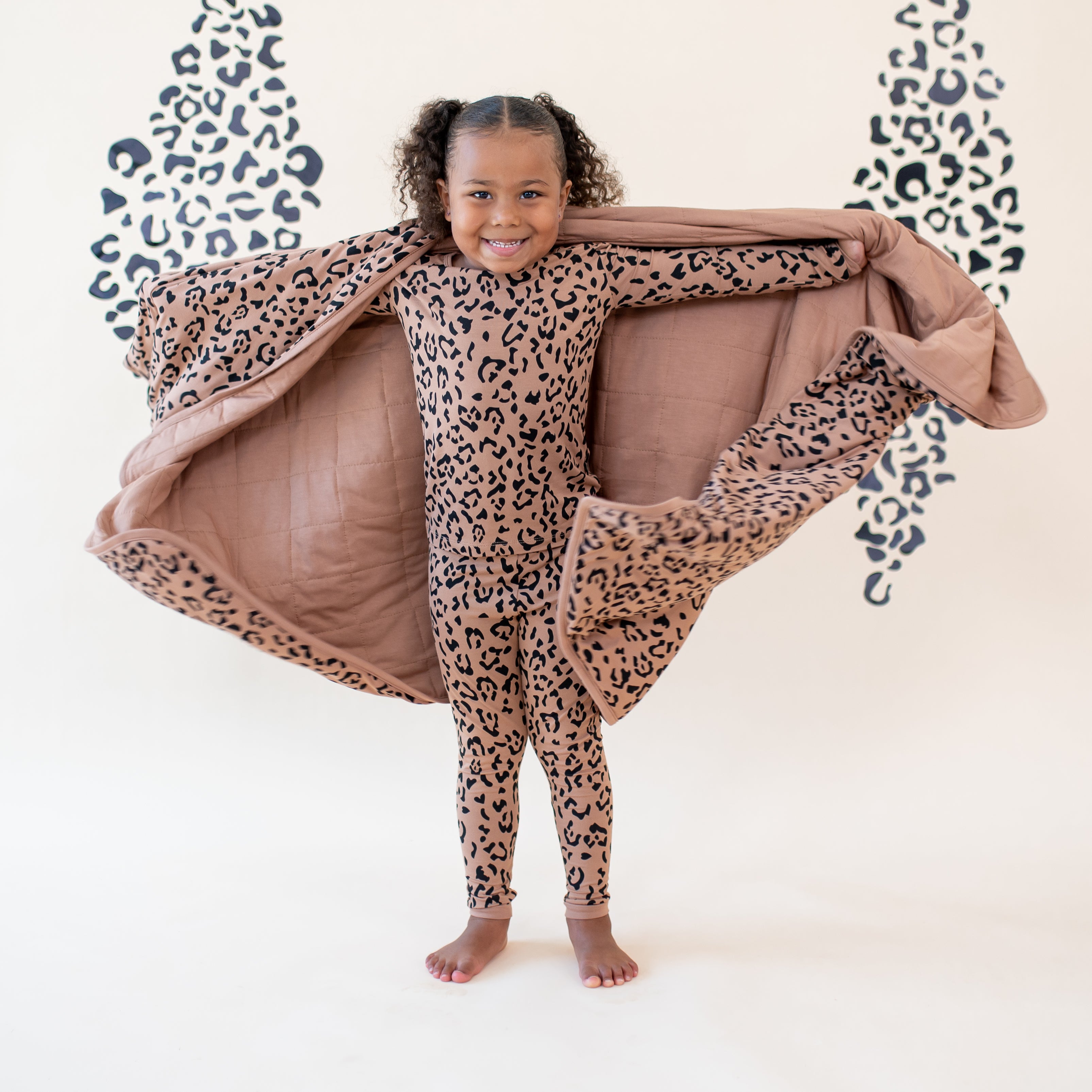 Smiling girl standing with her arms out holding a latte leopard toddler blanket wearing Long Sleeve Pajamas in Latte Leopard