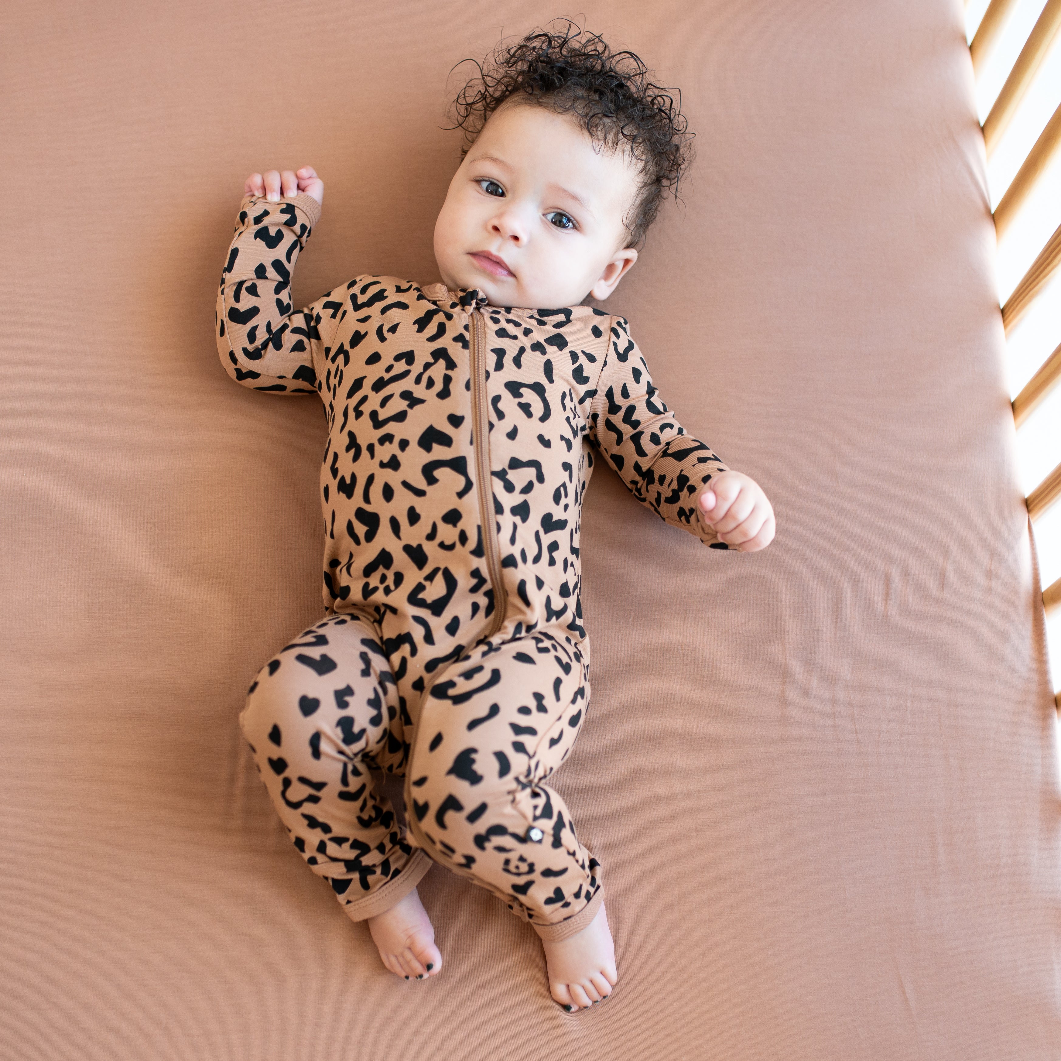 Infant laying in a crib on a latte crib sheet wearing the Zippered Romper in Latte Leopard