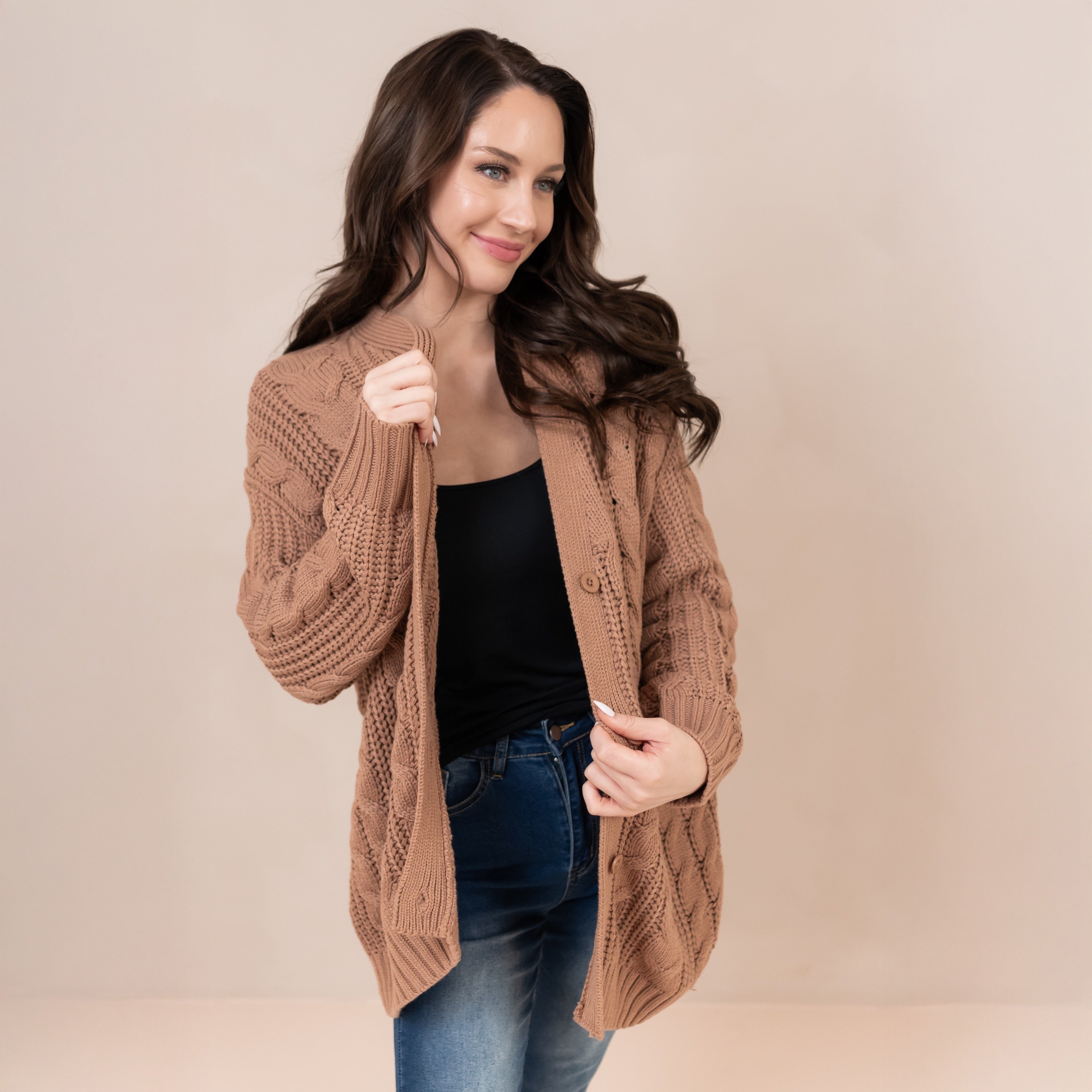 Female model standing wearing the Cable Knit Adult Cardigan in Latte holding the openings of the cardigan in both of her hands