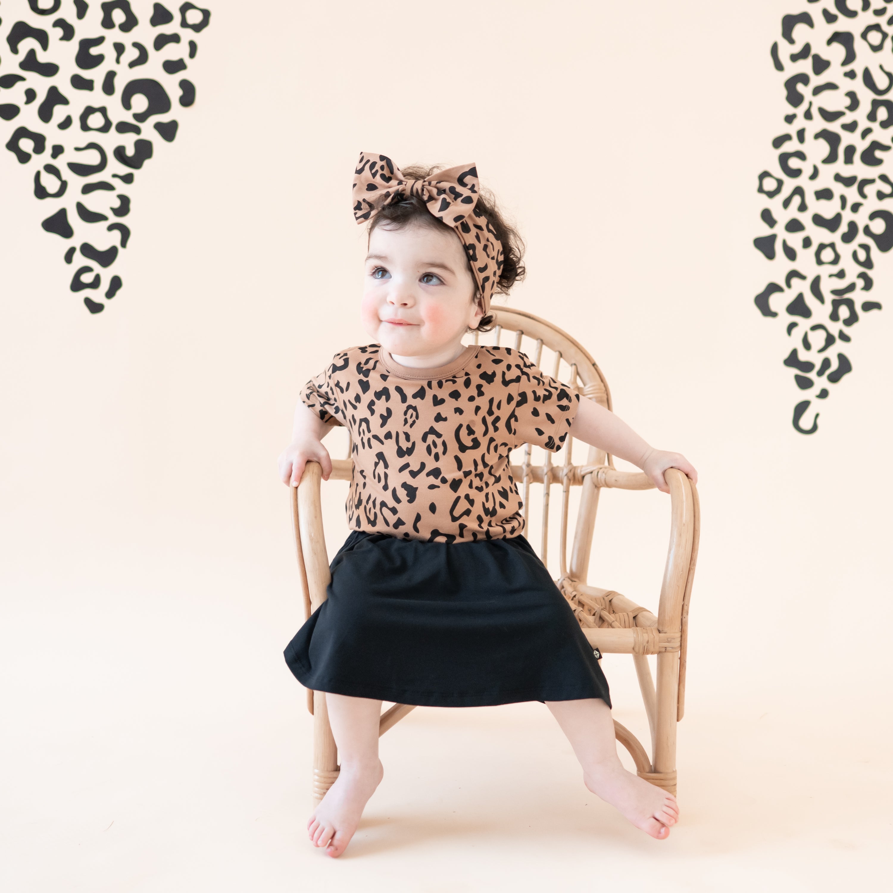 Toddler sitting on a wicker chair wearing the Bow Headband in Latte Leopard in the matching basic tee and midnight skort