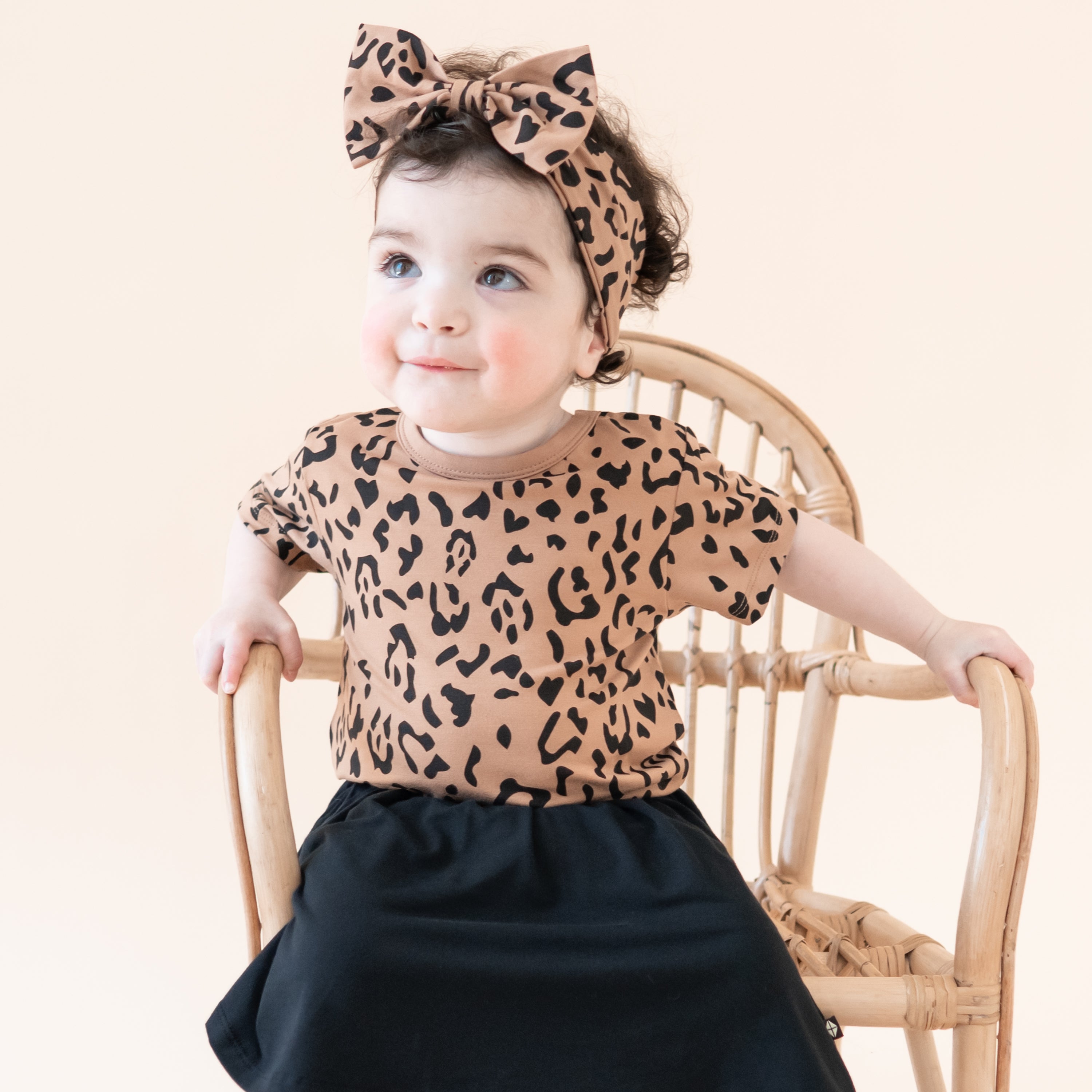 Toddler sitting in a wicker chair looking up wearing the Toddler Basic Tee in Latte Leopard with matching bow headband and midnight skort
