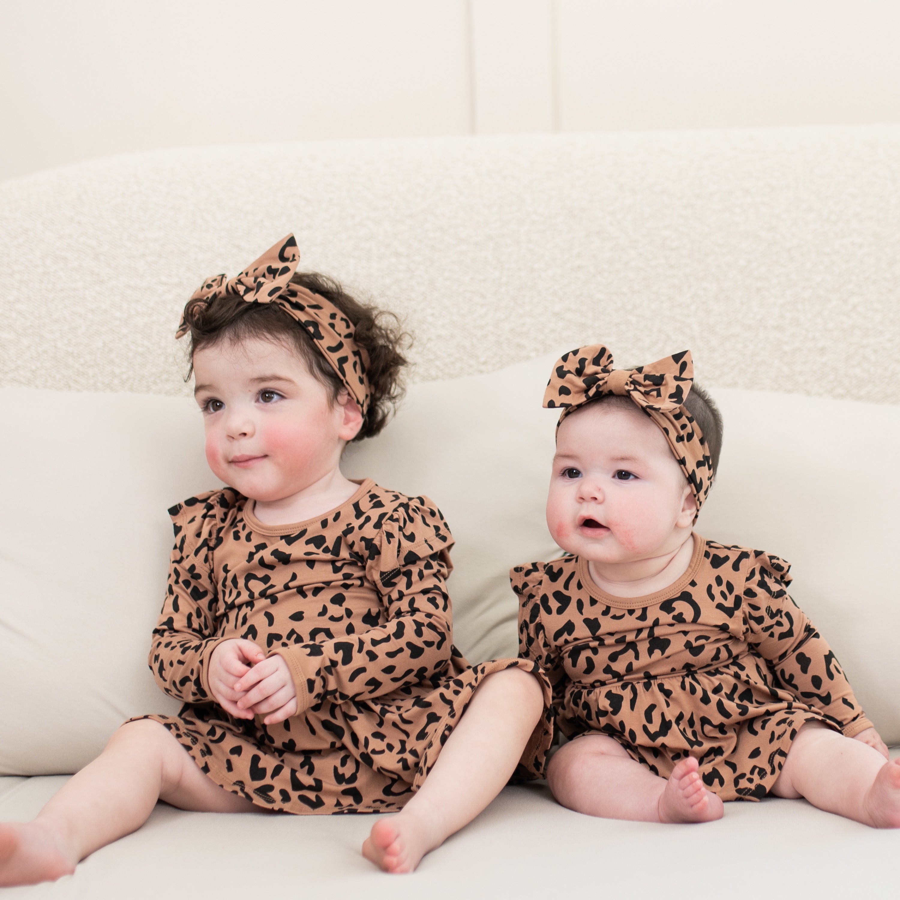 A toddler and infant sitting beside one another on the couch matching in a Long Sleeve Twirl Bodysuit Dress in Latte Leopard and matching bow headband