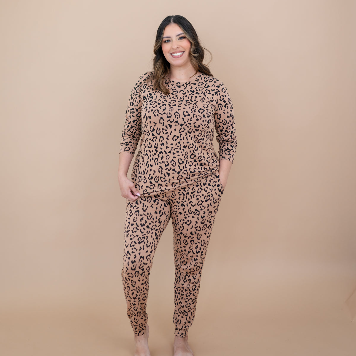 Smiling female standing with one hand in the pants pocket of the Women's Jogger Pajama Set in Latte Leopard in front of a taupe backdrop