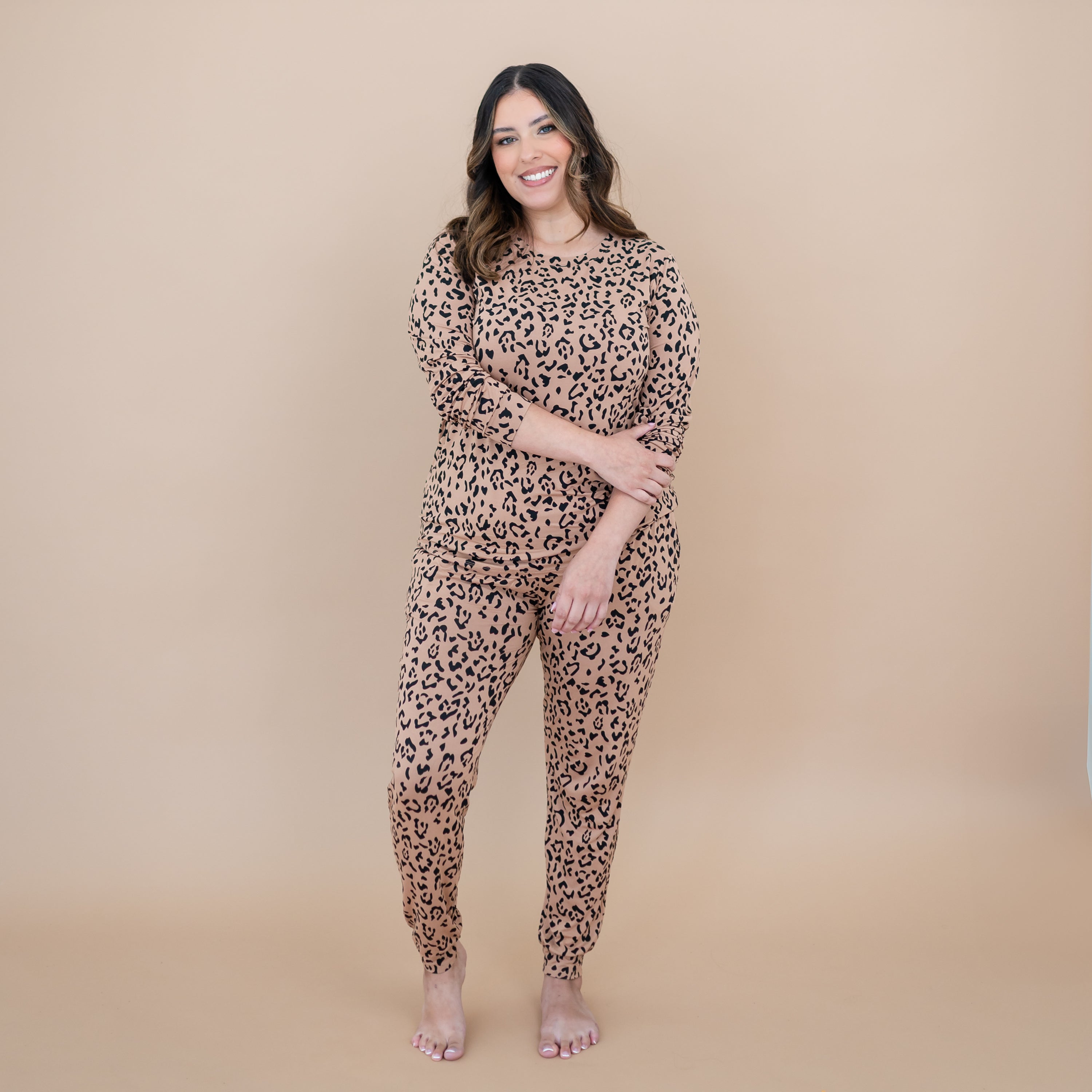 Smiling model standing with one hand holding the opposite arm wearing the Women's Jogger Pajama Set in Latte Leopard in front of a taupe background