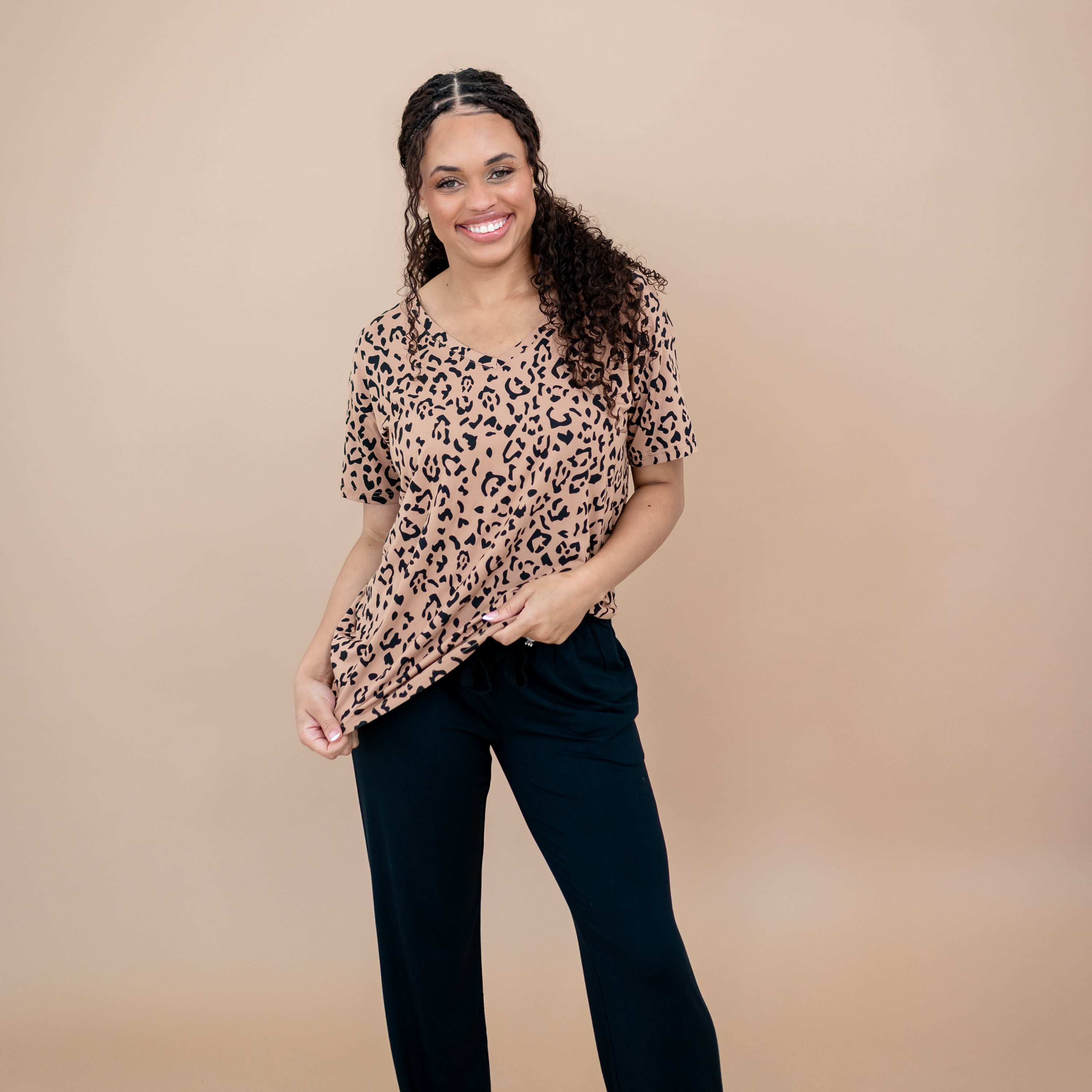 Smiling model standing in front of a taupe backdrop holding the bottom hem of the Women's Relaxed Fit V-Neck in Latte Leopard paired with black pants
