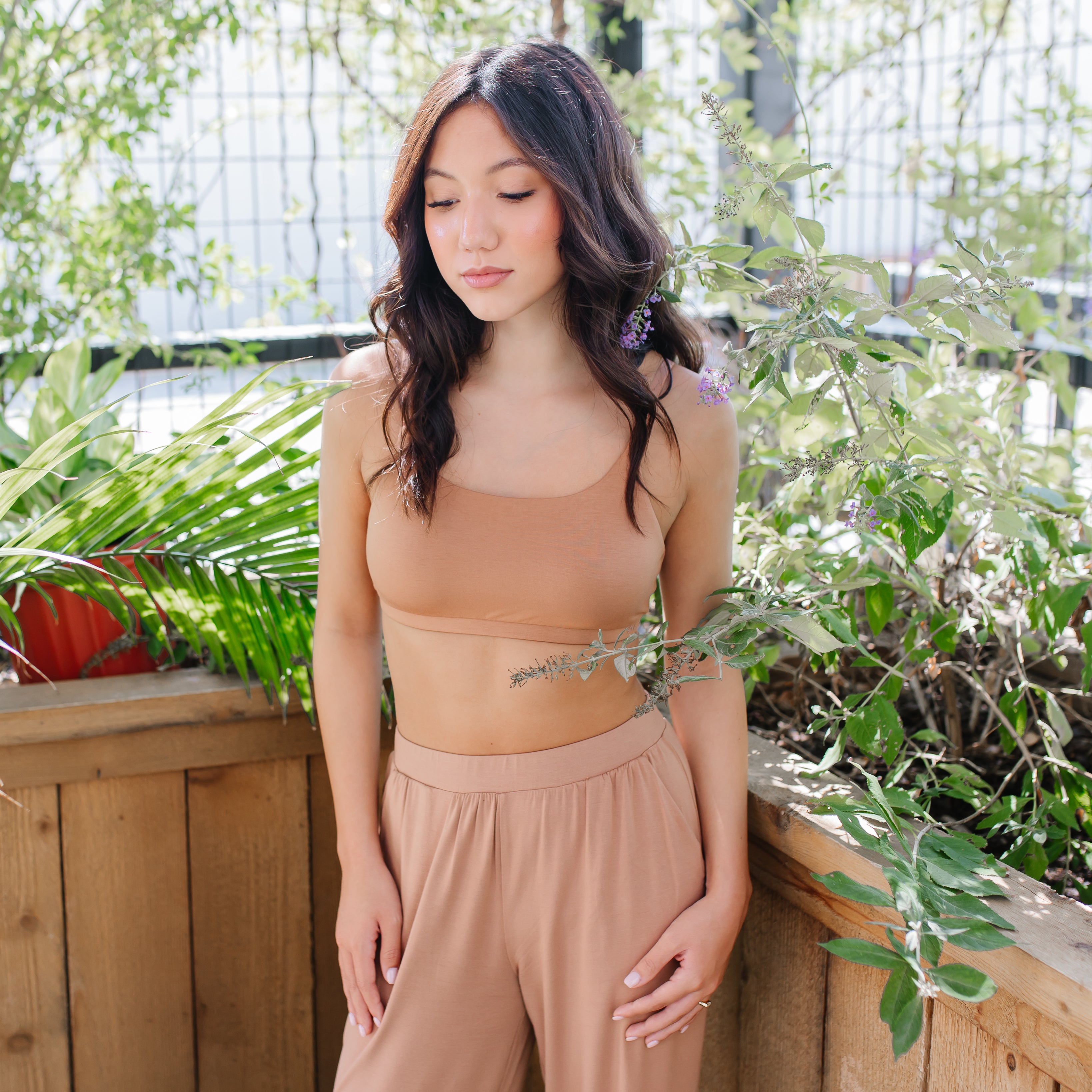 Female model standing in front of a wooden planter with greenery in it wearing the Women's Scoop Neck Bralette in Latte and light beige pants