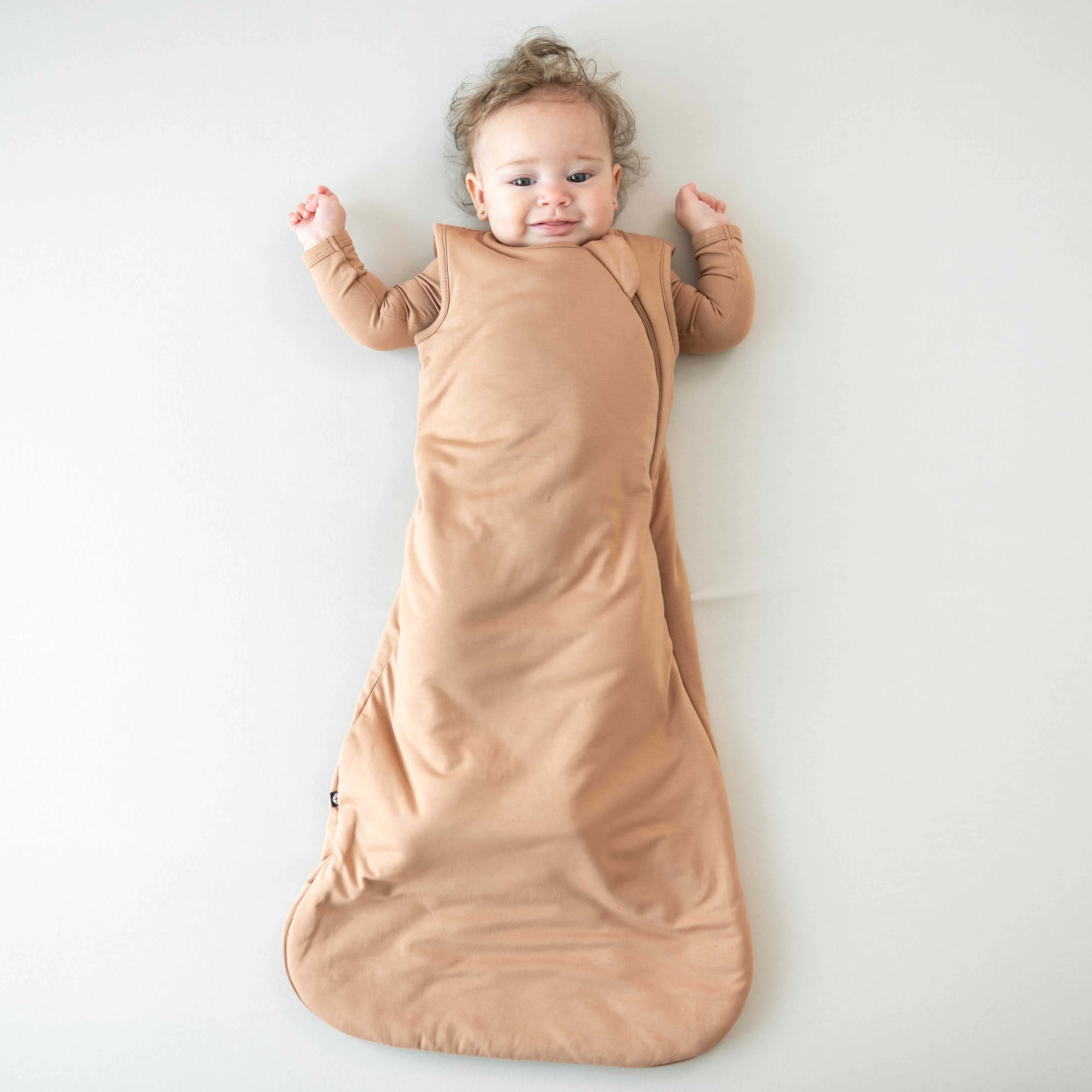 Infant laying down wearing the Sleep Bag in Latte 1.0 with matching long sleeve romper on underneath