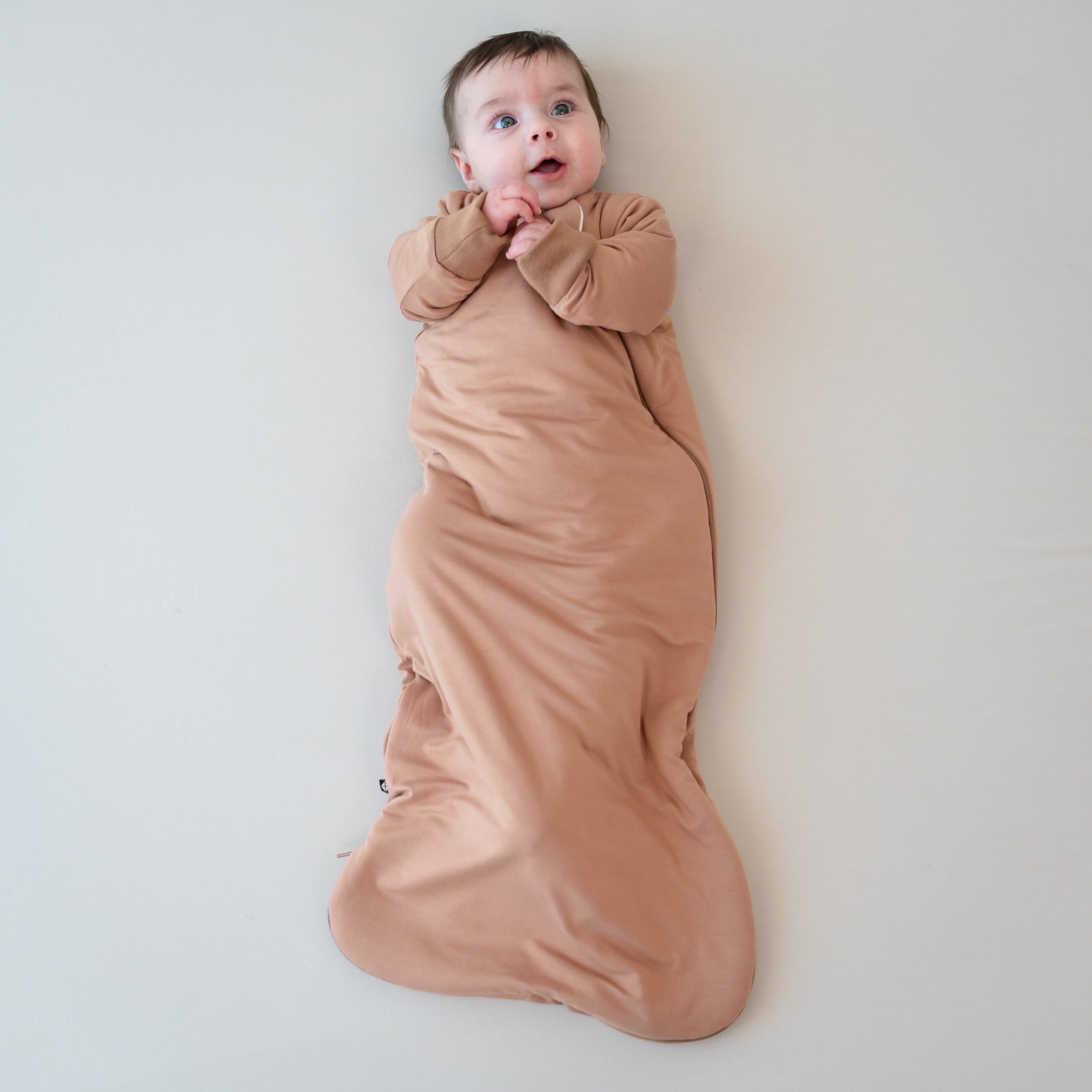 Infant laying down with hands clasped together wearing the Slumber Bag in Latte