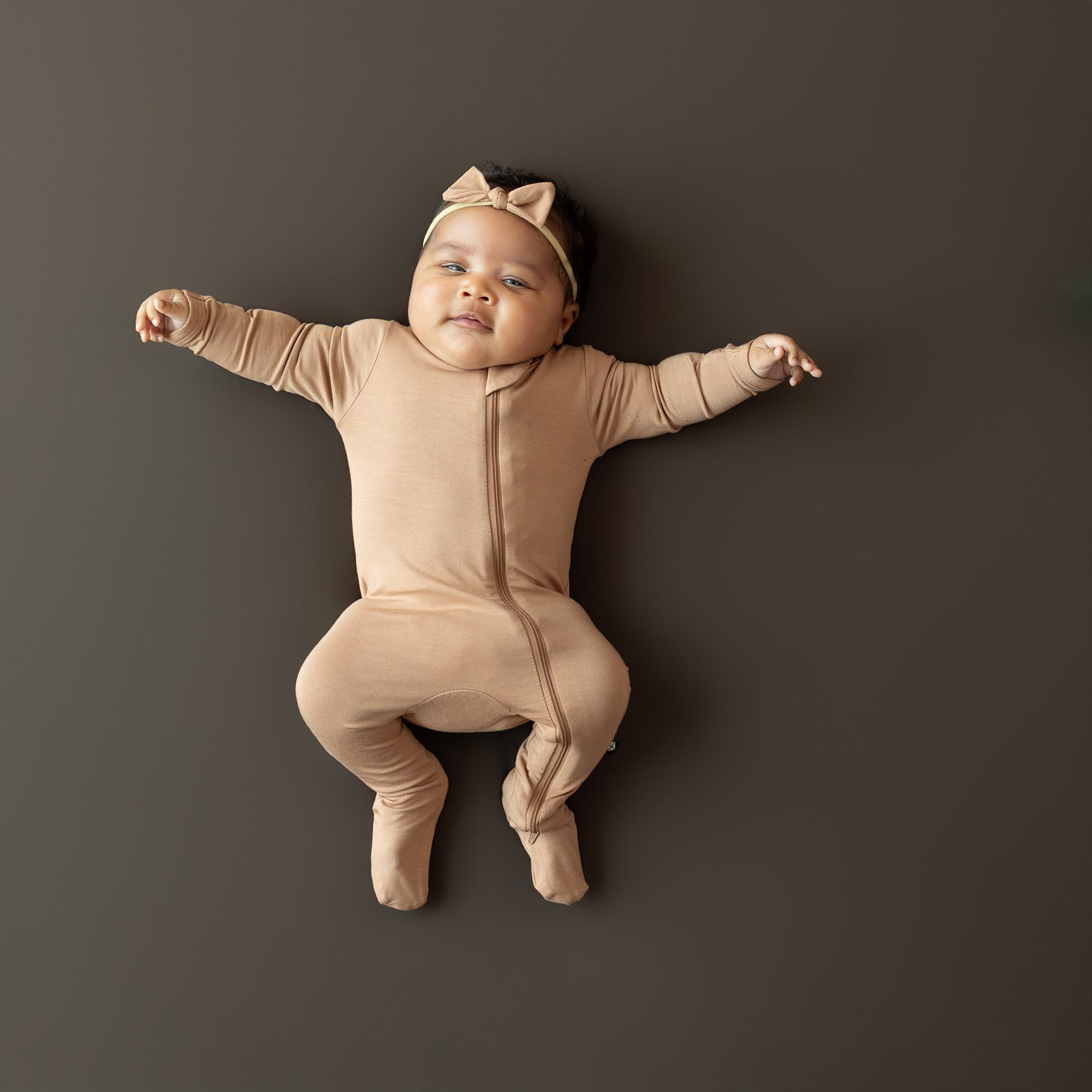 Baby in bamboo Zippered Footie in Latte and tiny bow lying on a dark background