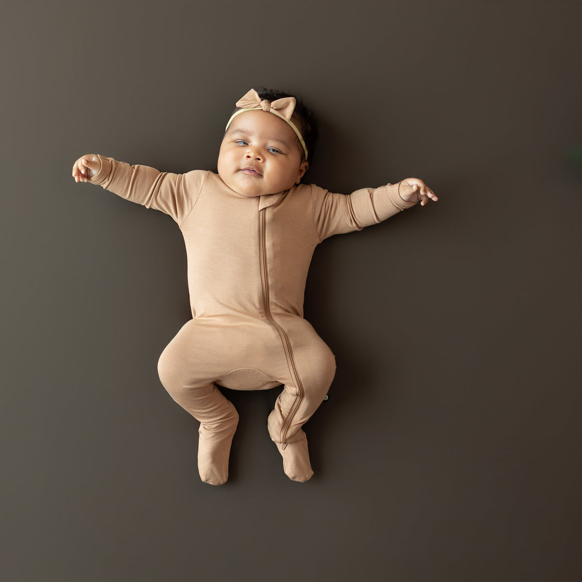 Baby in bamboo Zippered Footie in Latte and tiny bow lying on a dark background