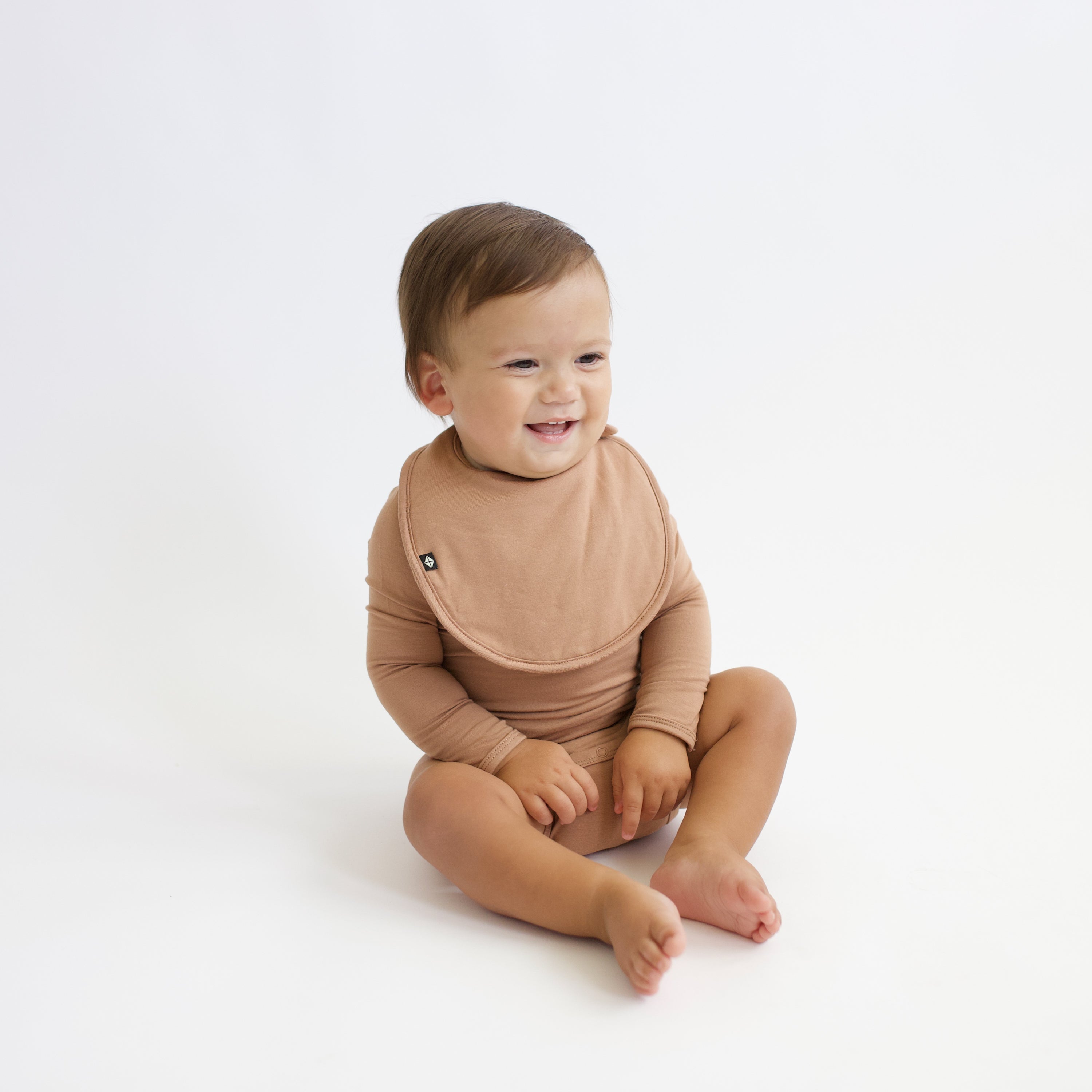 Smiling infant sitting wearing the Cushy Bib in Latte with the matching long sleeve bodysuit