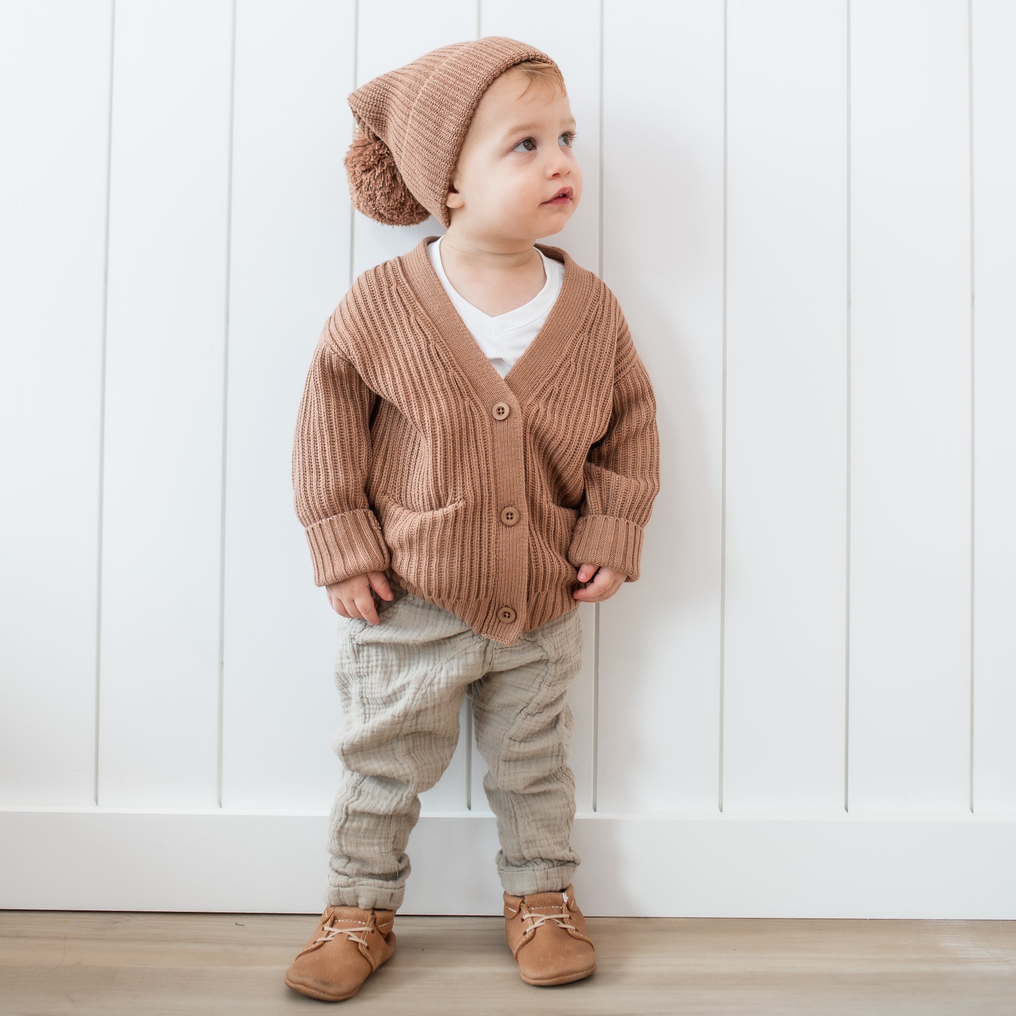 Toddler standing in front of a white paneled wall wearing the Chunky Knit Oversized Cardigan in Latte and matching chunky knit pom beanie