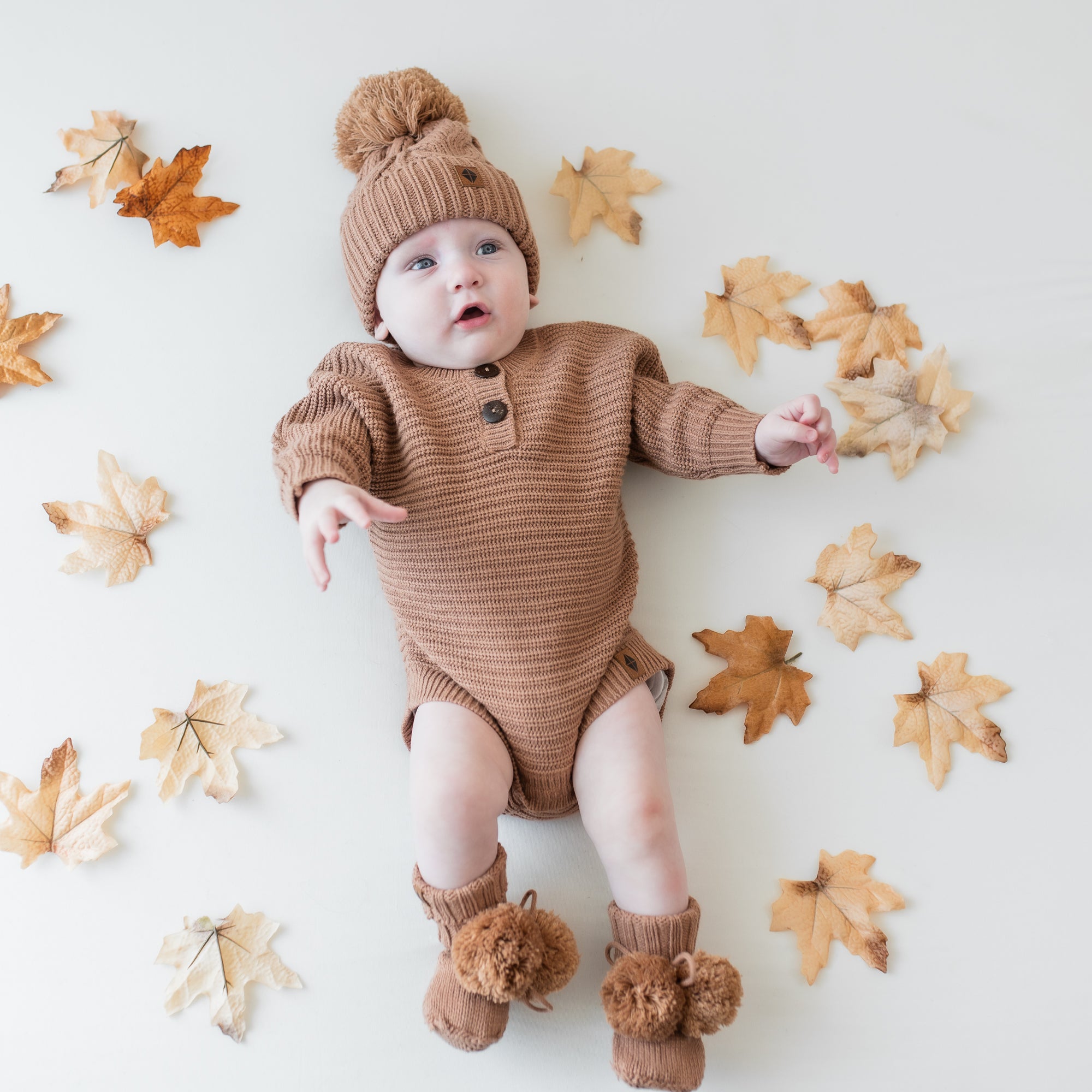 Infant wearing the Chunky Knit Romper in Latte with matching knit booties and chunky cable knit beanie surrounded by prop leaves