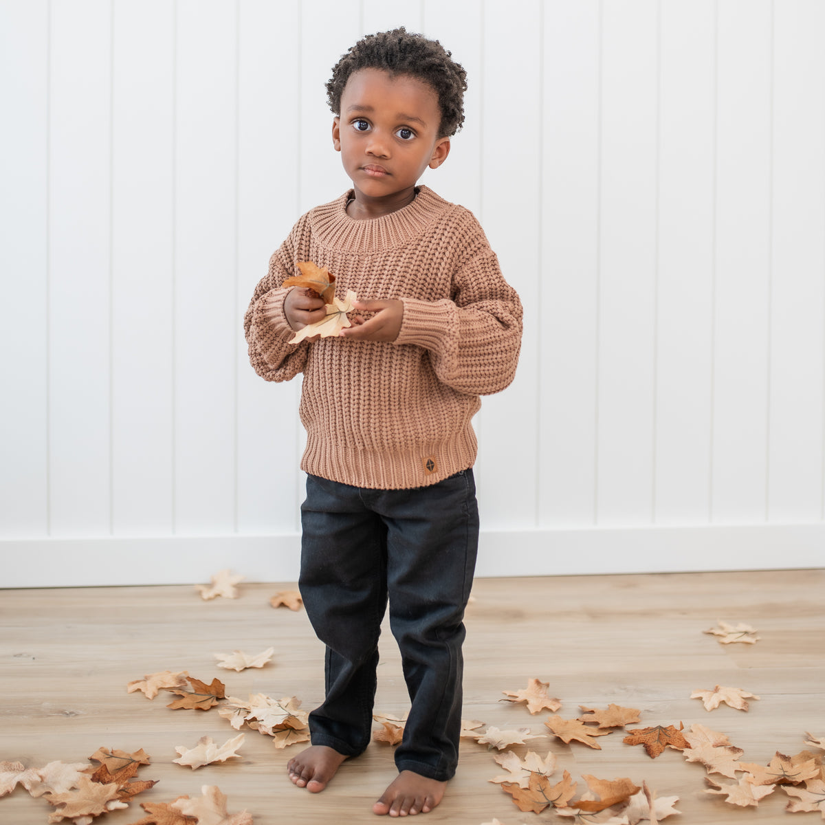 Young boy wearing the Chunky Knit Sweater in Latte with black jeans standing in front of a white paneled wall holding leaves