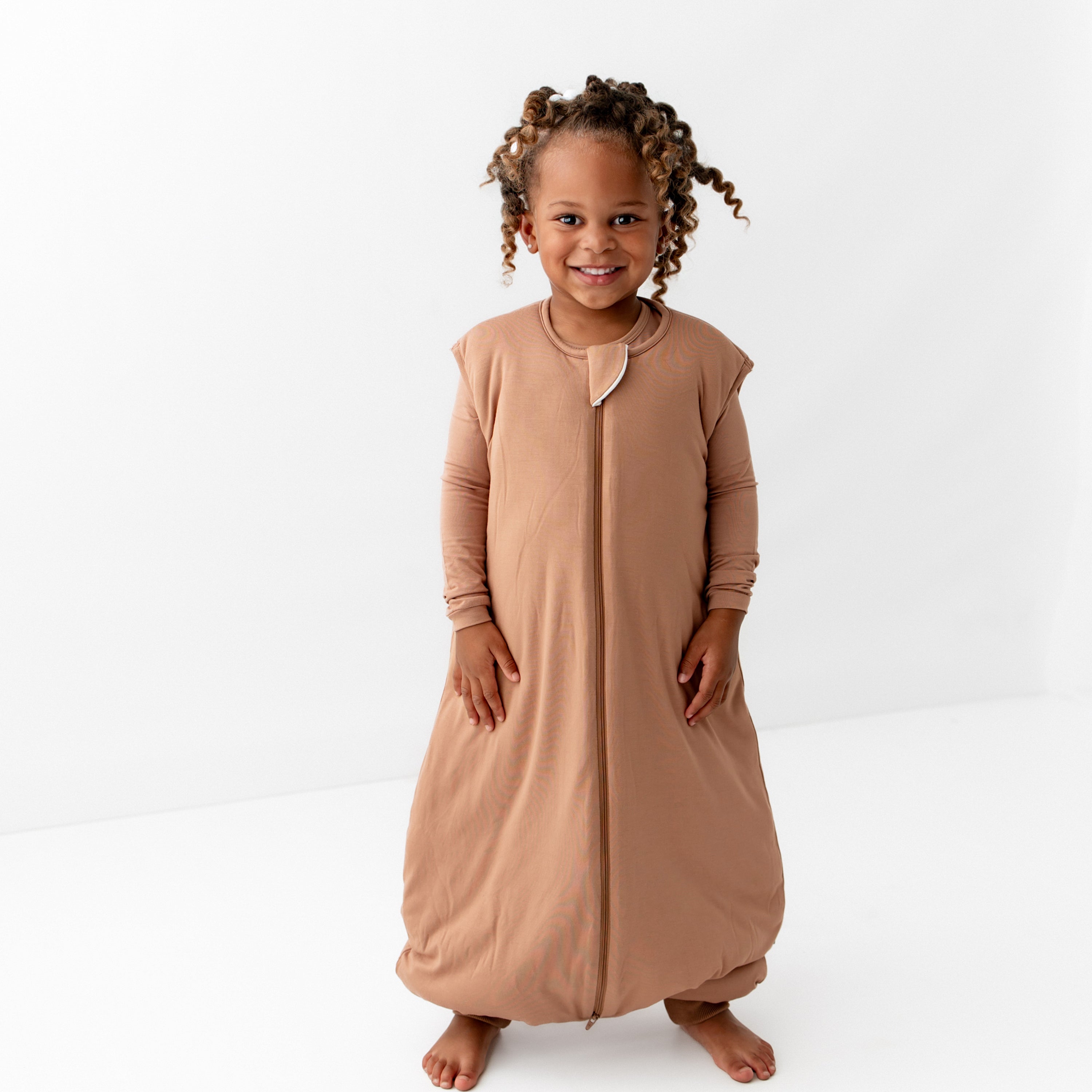 Toddler standing with their hands at their side in the Sleep Bag Walker in Latte 2.5 with the matching long sleeve toddler pajamas on underneath