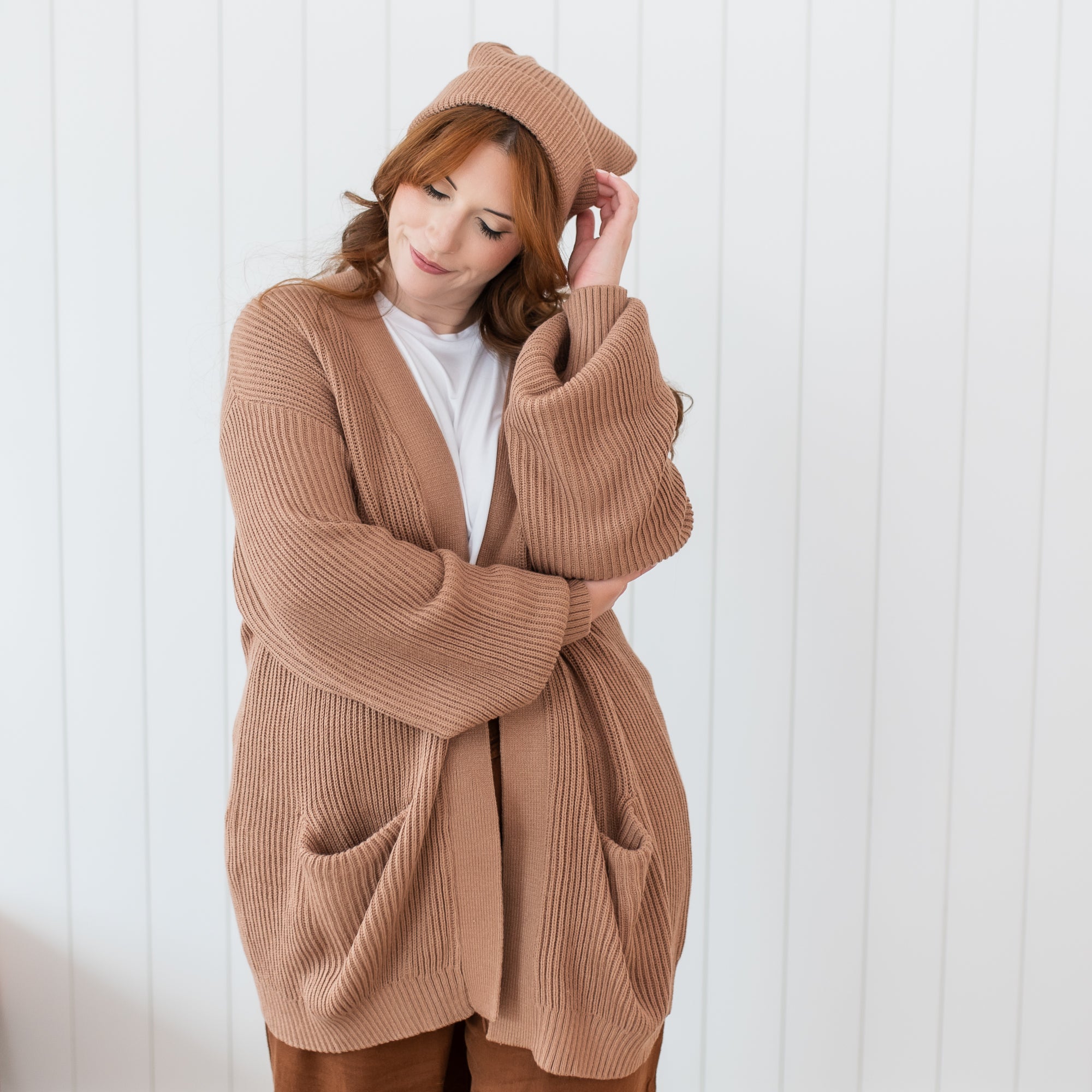 Female model standing in front of a white paneled wall wearing the Chunky Knit Women's Oversized Cardigan in Latte and matching chunky knit Adult pom beanie