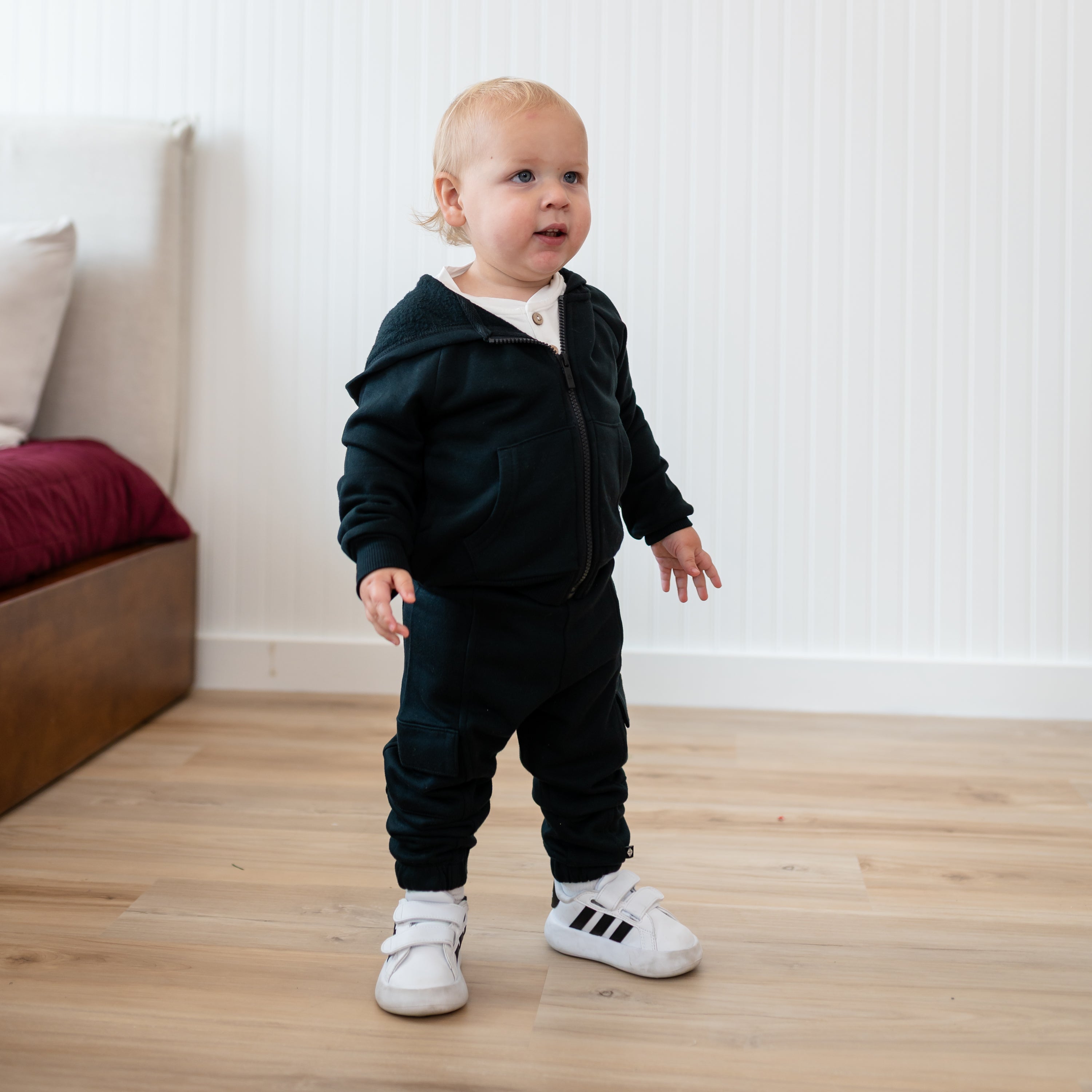 Young toddler standing beside a bed wearing the Fleece Cargo Pants in Midnight paired with a matching fleece zip hoodie