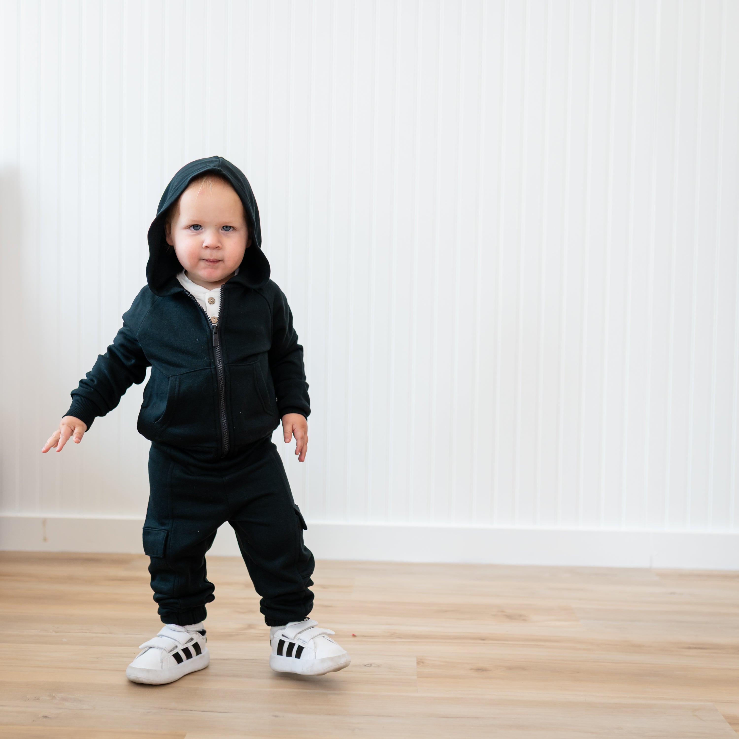 Young toddler standing in front of a white paneled wall wearing the Fleece Zip Up Hoodie in Midnight and matching fleece cargo pant