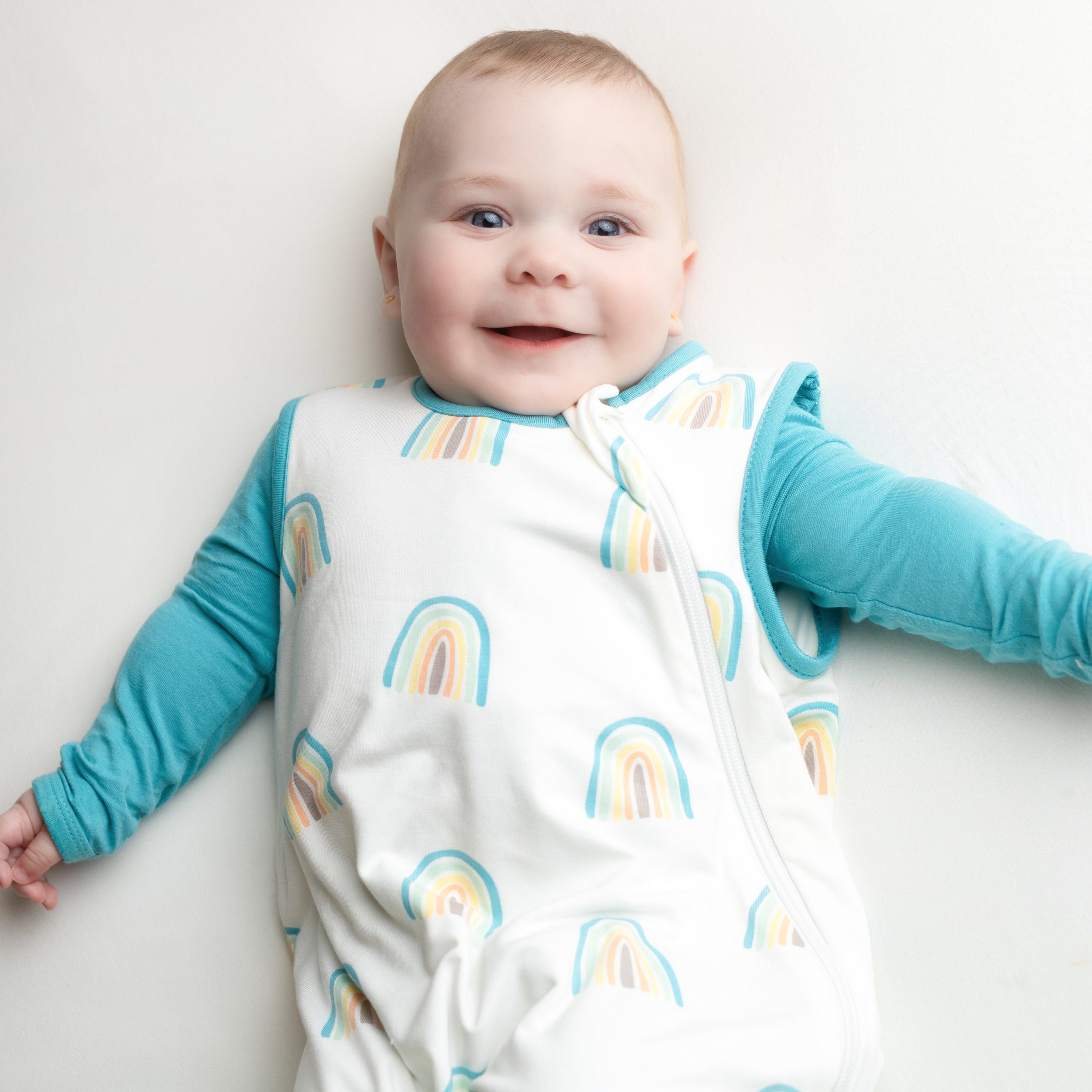 Smiling baby wearing the Sleep Bag in Makai Rainbow 0.5 with a Makai Long Sleeve Romper underneath