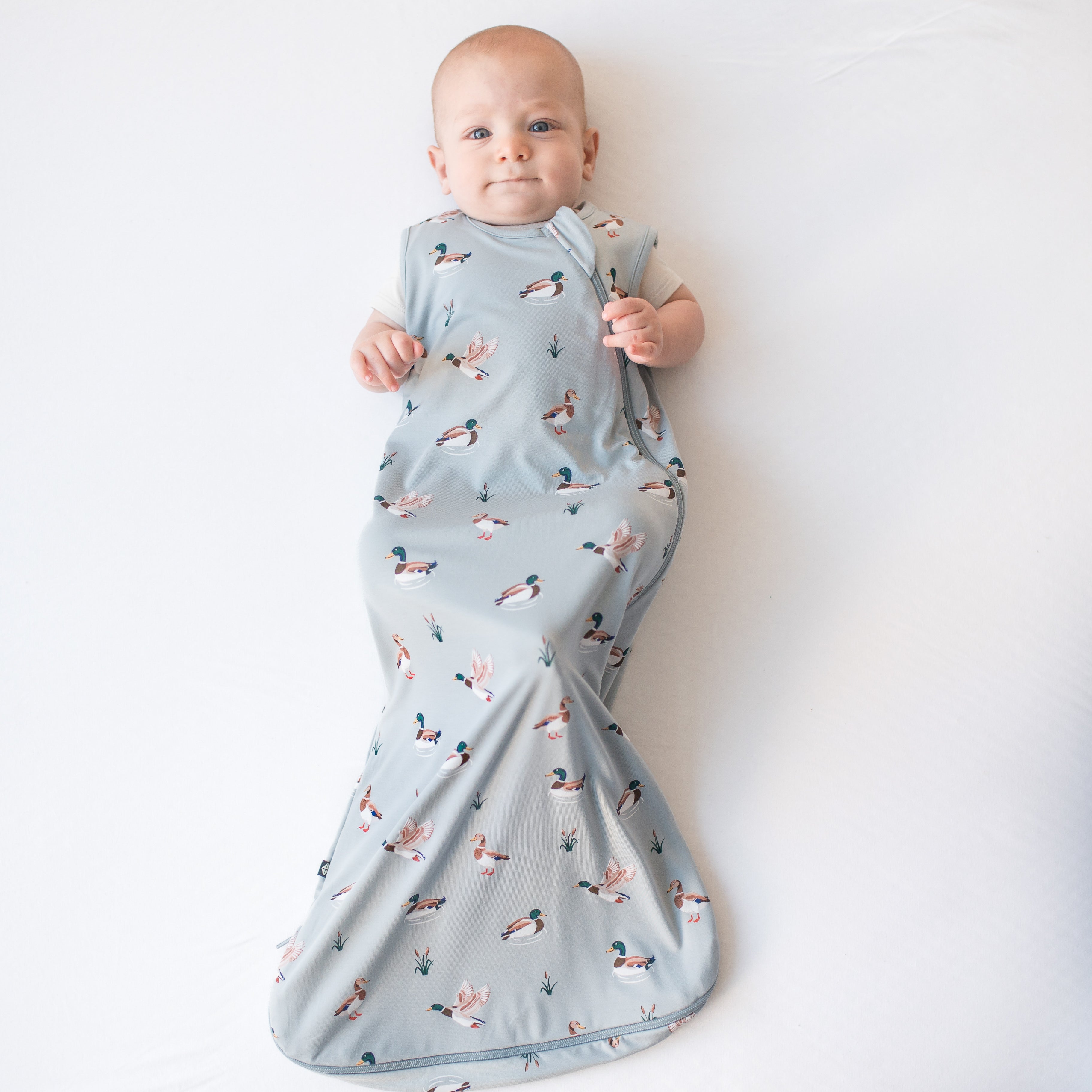 Infant laying on a white surface wearing the Sleep Bag in Mallard