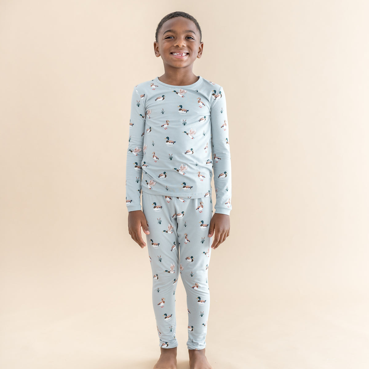 Young smiling boy wearing the Long Sleeve Pajamas in Mallard Duck in front of a light beige background