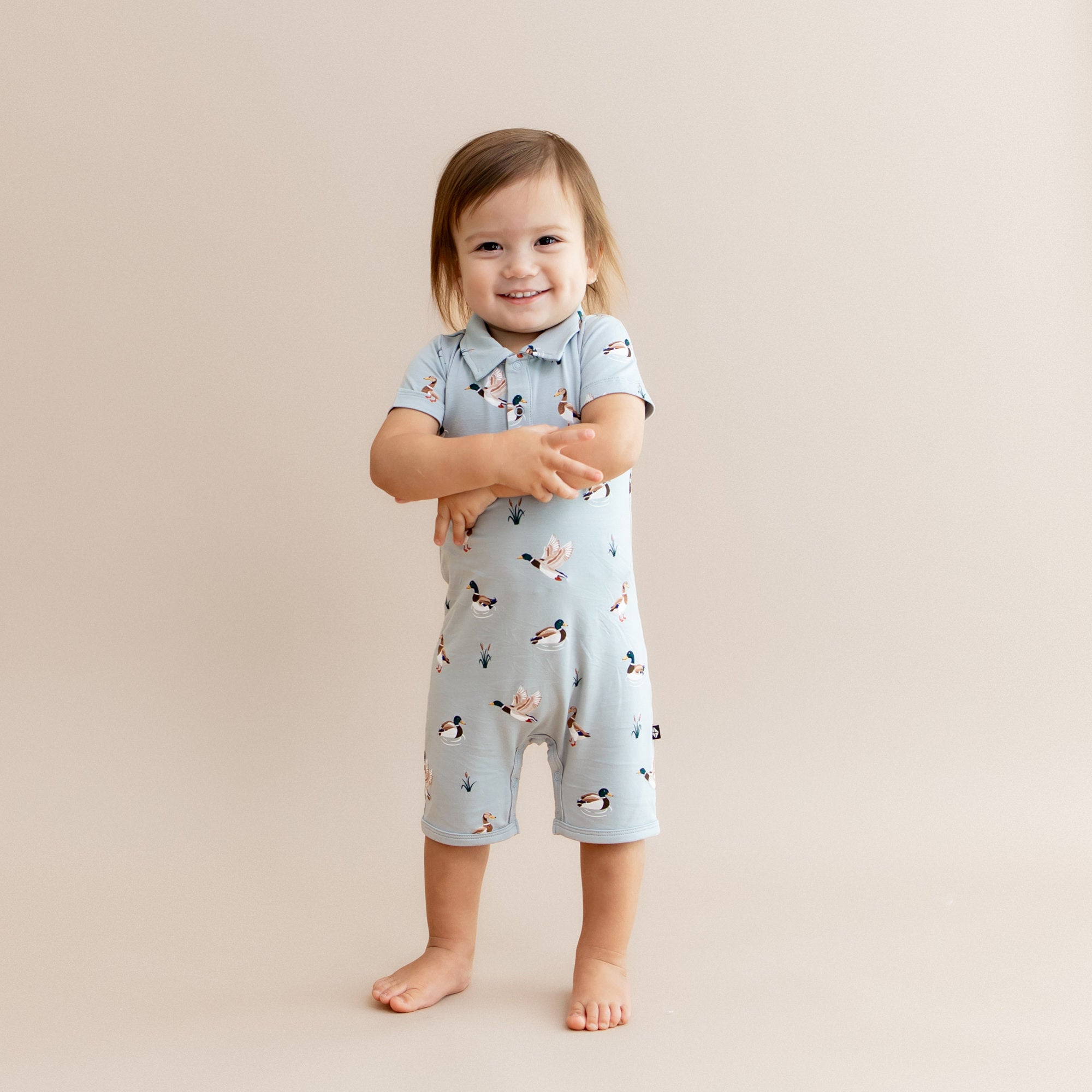 Young smiling toddler standing in front of a light beige background wearing the Polo Shortall in Mallard Duck