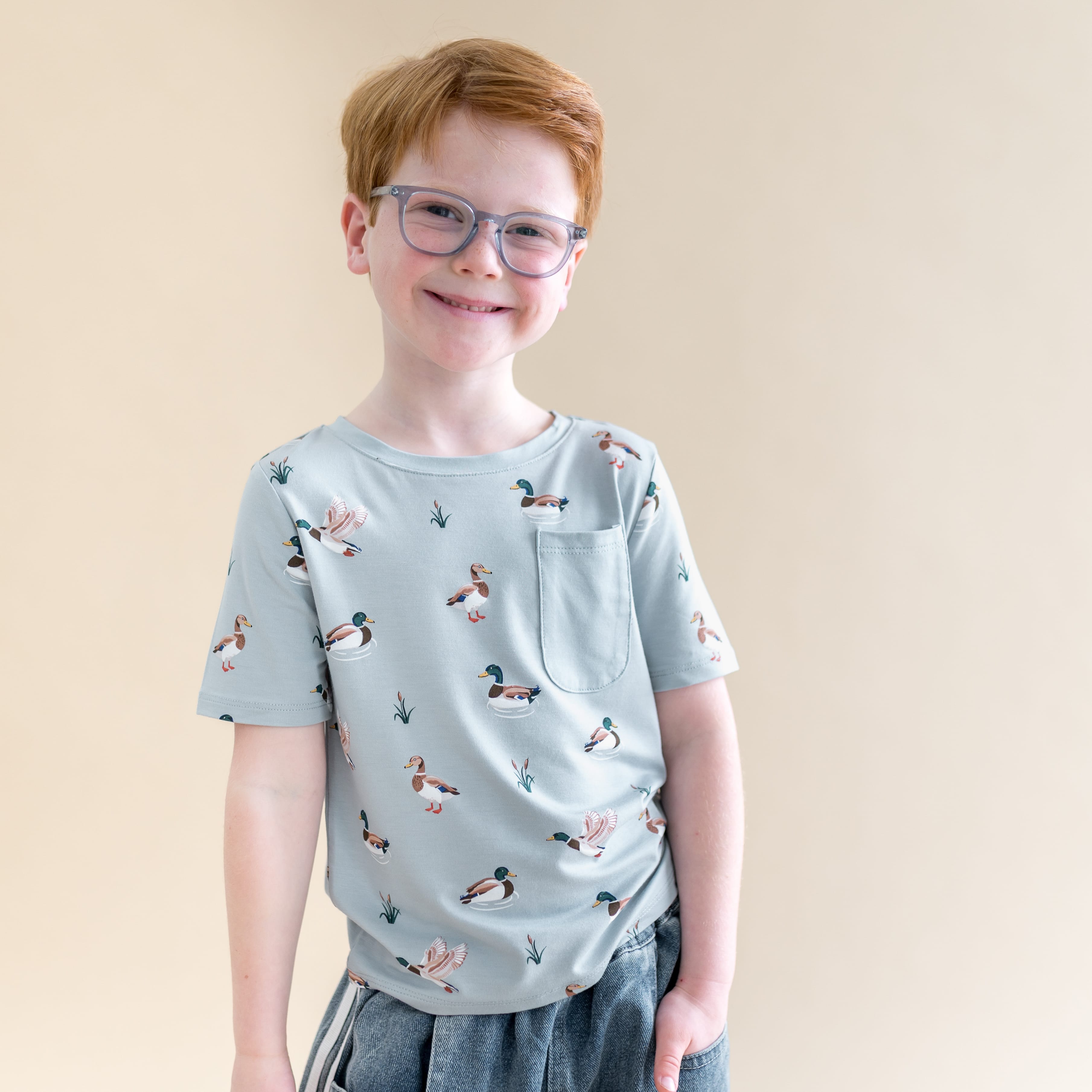 Smiling boy wearing the Toddler Crew Neck Tee in Mallard Duck standing in front of a light taupe background