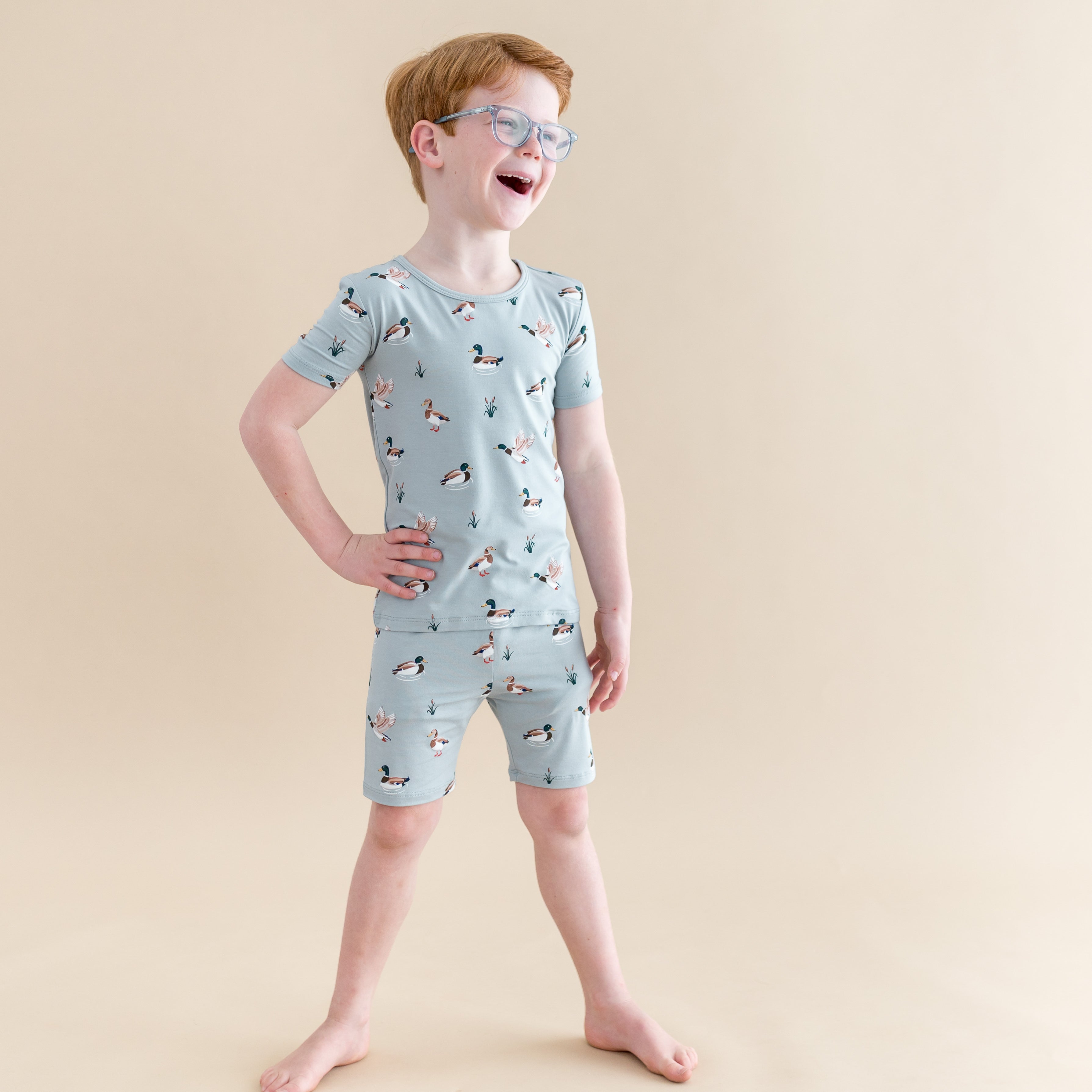 Smiling boy modeling the Short Sleeve Pajamas in Mallard Duck standing in front of a light taupe background