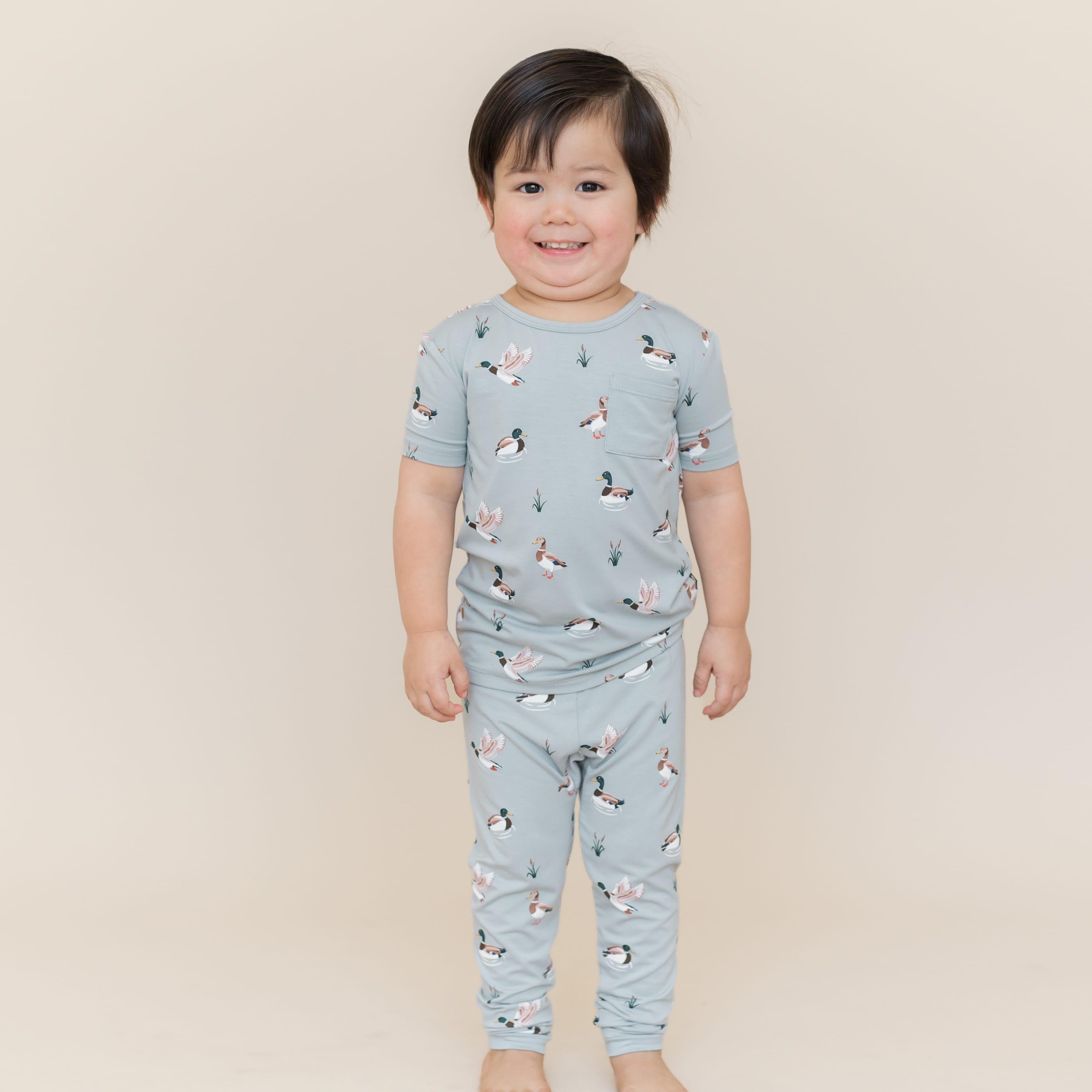 Smiling boy standing in front of a light taupe background wearing the Short Sleeve with Pants Pajamas in Mallard Duck