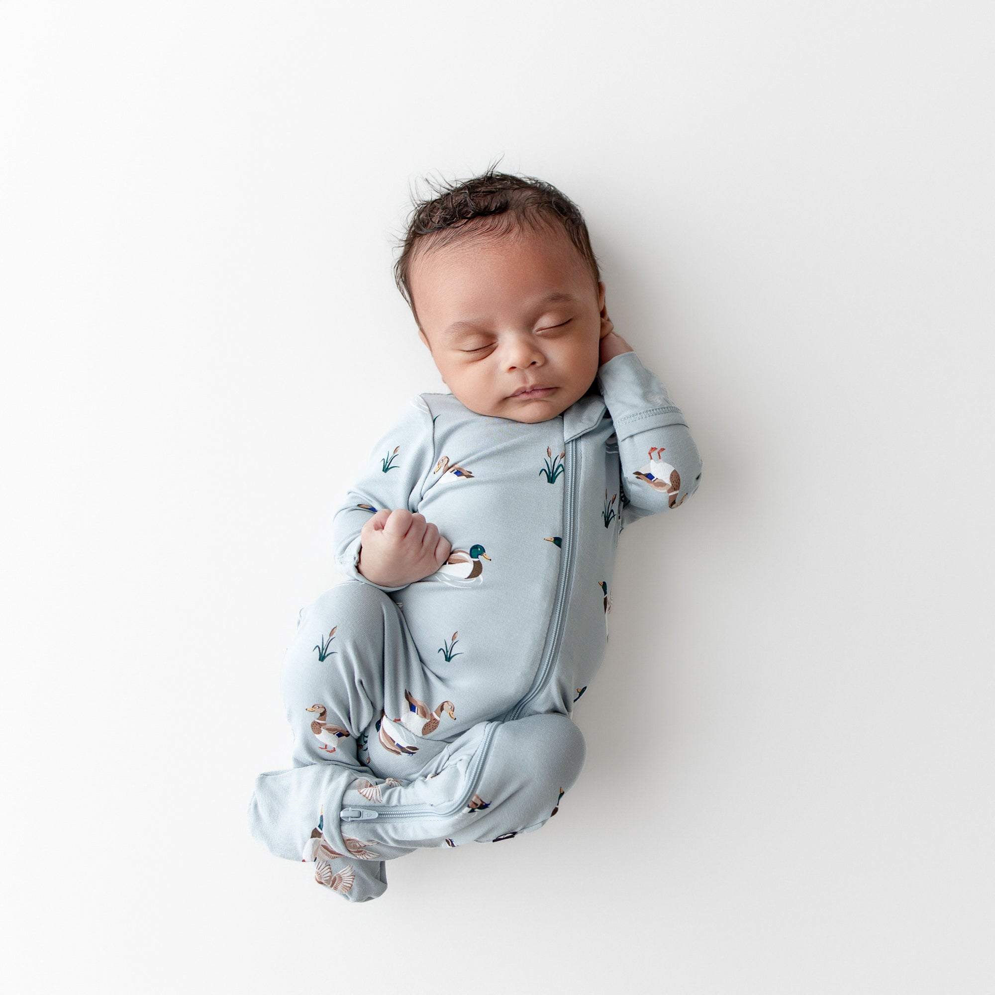 Sleeping newborn wearing the Zippered Footie in Mallard Duck laying on a white surface