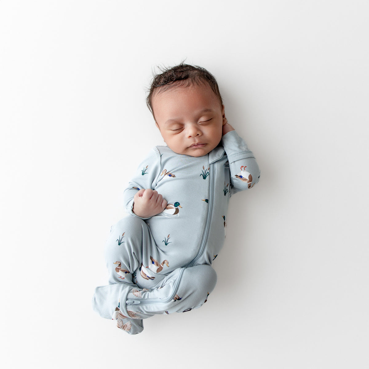 Sleeping newborn wearing the Zippered Footie in Mallard Duck laying on a white surface